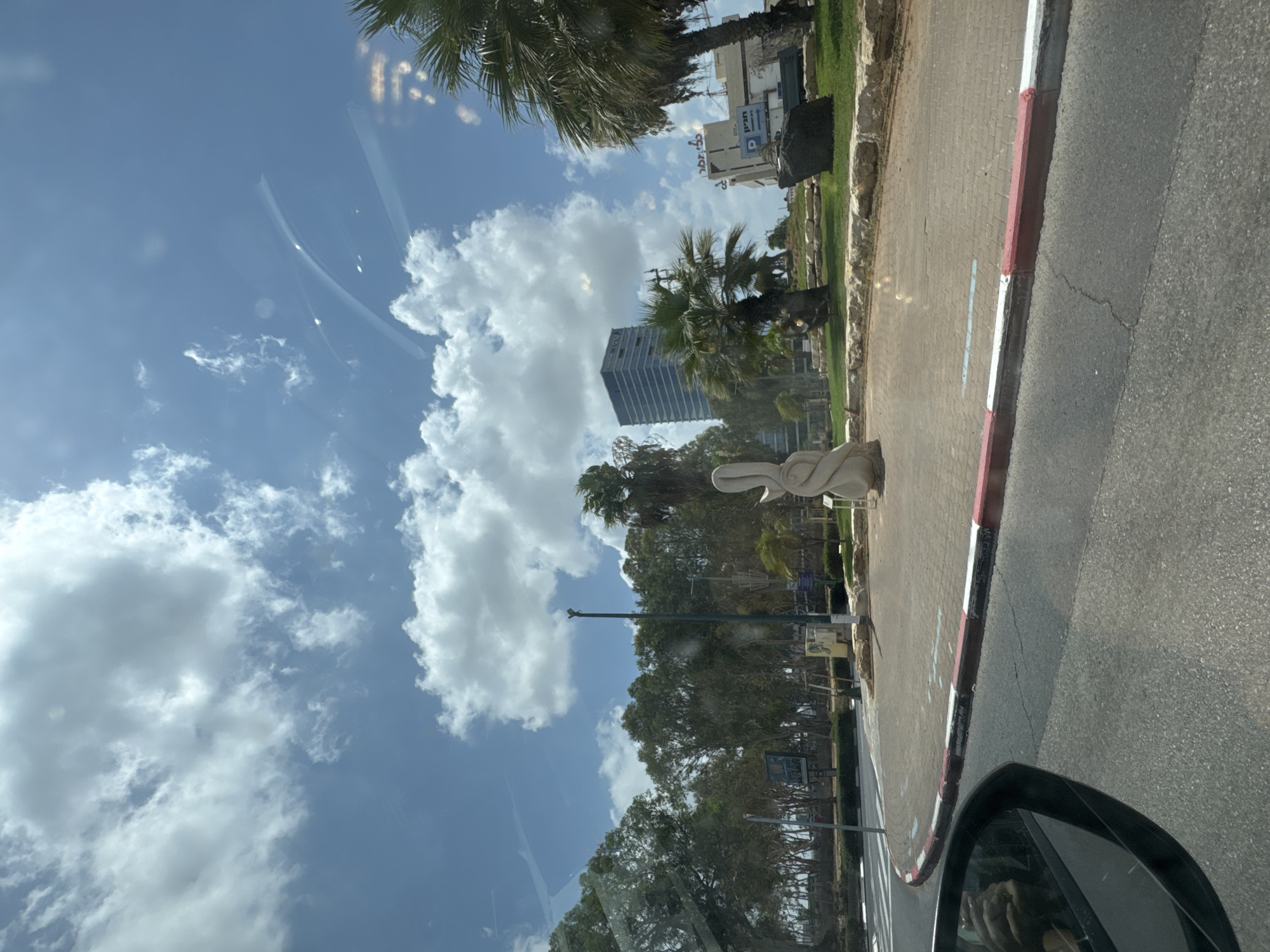 The image, captured from a vehicle in Petah Tikva, Israel, depicts a public space on a clear day with abundant white clouds scattered across a blue sky. In the foreground, the edge of an asphalt road is visible, with a curb marked by red and white stripes. To the right of the road, a paved walkway with brick-like pavers is present, bordered by a low stone wall and green grass. A white sculpture, abstract and twisting, stands on the paved area. Behind the walkway, a park-like setting is established with various trees, including palm trees, and some small bushes. A tall, slender lamppost is centrally located. In the background, a modern, multi-story glass building is prominent, and further to the right, another building complex with signage is visible. Reflections are present on the window through which the photo was taken.