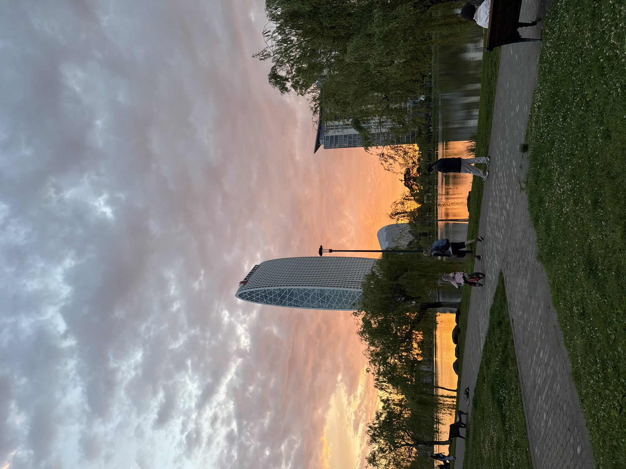 The image captures a park scene in Batumi, Georgia, at sunset. A body of water reflects the orange and pink hues of the sky, contrasting with the silhouetted trees lining its banks. A paved walkway runs along the water's edge, where people are walking and cycling. In the background, modern architecture, including a distinctive oval-shaped building, punctuates the urban landscape. The sky above is filled with a mix of gray and orange clouds.