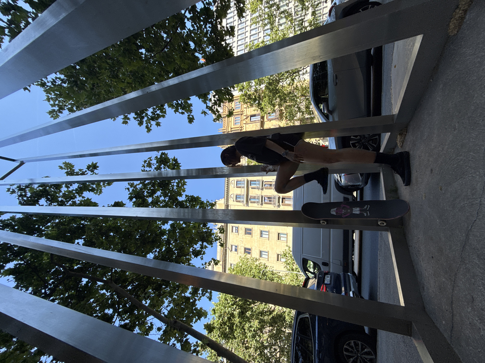 A person, wearing dark shorts and a dark t-shirt, is positioned between metal vertical beams and their skateboard is on the ground near them. Cars and trees are behind the metal structure. Buildings are in the background. The photo was taken outside in Barcelona, Spain, as evidenced by the license plates of the vehicles and architectural style of the buildings.