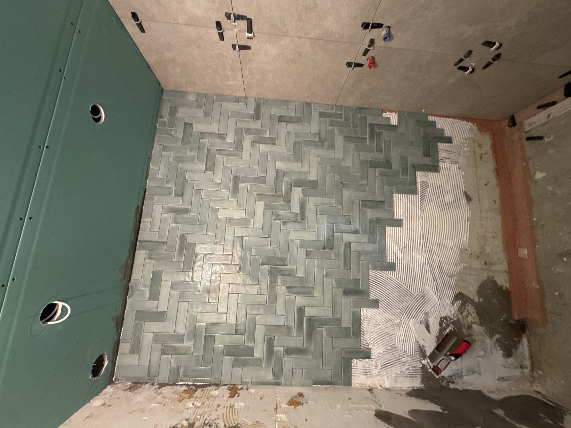 This image depicts an indoor space, likely a bathroom or wet room, currently under construction or renovation. The floor is prominently featured, with a significant portion covered in rectangular, light blue-grey tiles arranged in a herringbone pattern. These tiles exhibit variations in shade and potentially a slightly iridescent or handmade finish.
To the right of the tiled area, white tile adhesive is freshly spread on the bare subfloor, showing distinct, curved trowel marks, indicating that tiling is an ongoing activity. A red and black notched trowel rests on this adhesive, ready for use. The untiled sections of the floor appear to be bare concrete or a similar subfloor material, with some scattered debris.
The left wall is clad in green waterproof drywall or cement board, characteristic of wet area construction. This wall features several circular cutouts, presumably for plumbing pipes or electrical outlets. Visible screws are present along the edges of the board. The ceiling, and possibly the upper section of the back wall, are covered with larger, light beige-grey tiles. Protruding from these upper tiles are several black clips or ties, along with a red and a blue pipe cap, which suggest plumbing connections for fixtures like a shower or faucet. Tile leveling clips are also visible between the ceiling tiles. A section of the right wall, near the floor, appears to be an older surface, possibly concrete, treated with a reddish-brown primer or waterproofing layer.
The image captures a moment in time during a tiling project, showcasing the progression of work. No people or text are visible in the scene. The overall impression is one of careful and methodical installation in an interior setting, likely in Leiden, Netherlands.