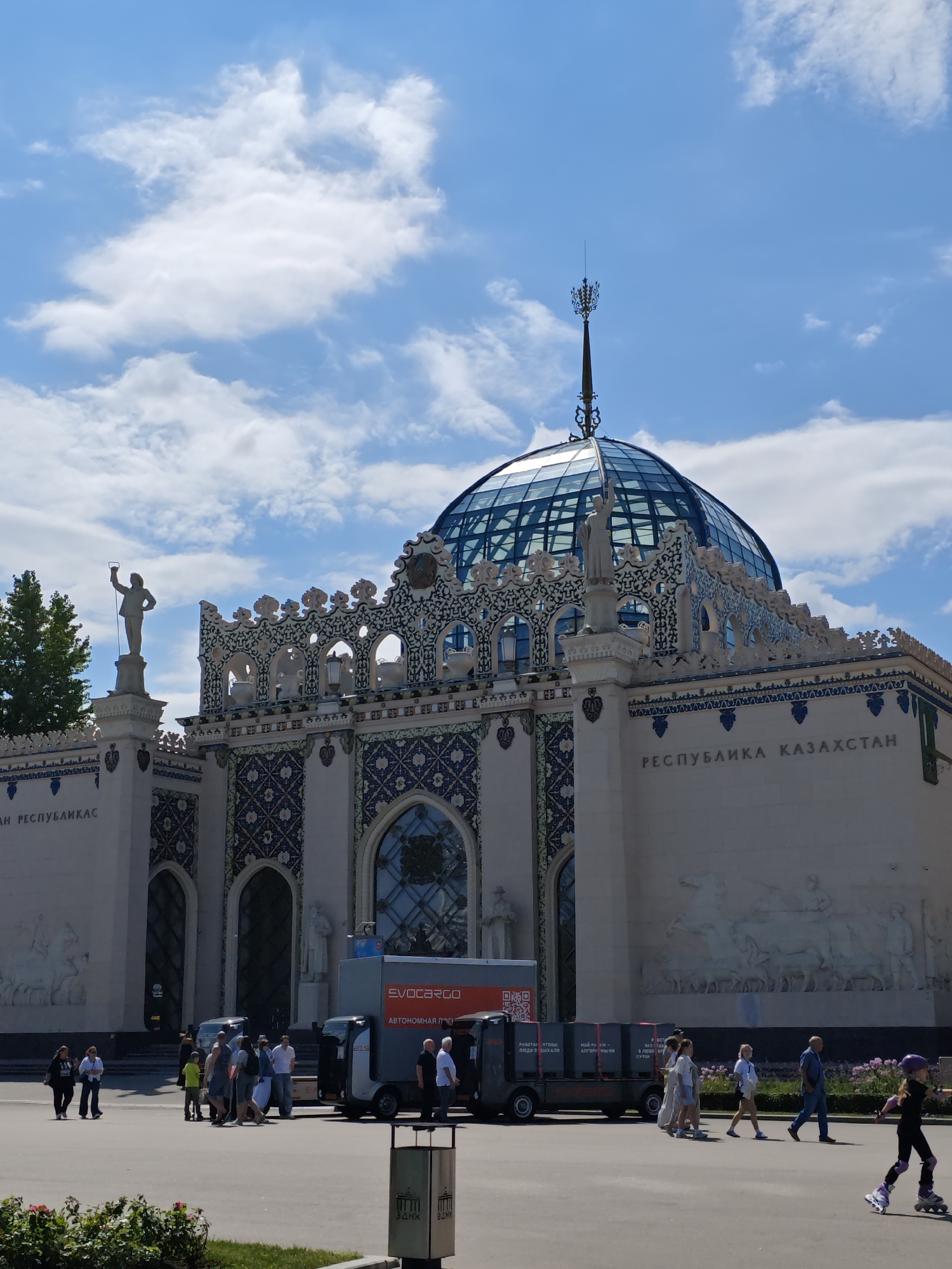 This is a photograph of the Kazakhstan Pavilion at VDNKh (Exhibition of Achievements of National Economy) in Moscow, Russia. The pavilion, built in 1939 and rebuilt in 1954, features a distinctive blue glass dome topped with a spire. The facade is adorned with intricate blue and white ornamental patterns, and bas-reliefs depicting rural life. On the left side of the pavilion, a statue of a worker holding a torch stands prominently. Several people are walking in front of the pavilion, and a truck with the "EVOCARGO" logo is parked nearby. The sky is blue with scattered white clouds. The surrounding area includes green trees and a grassy area with flowers in the foreground.
