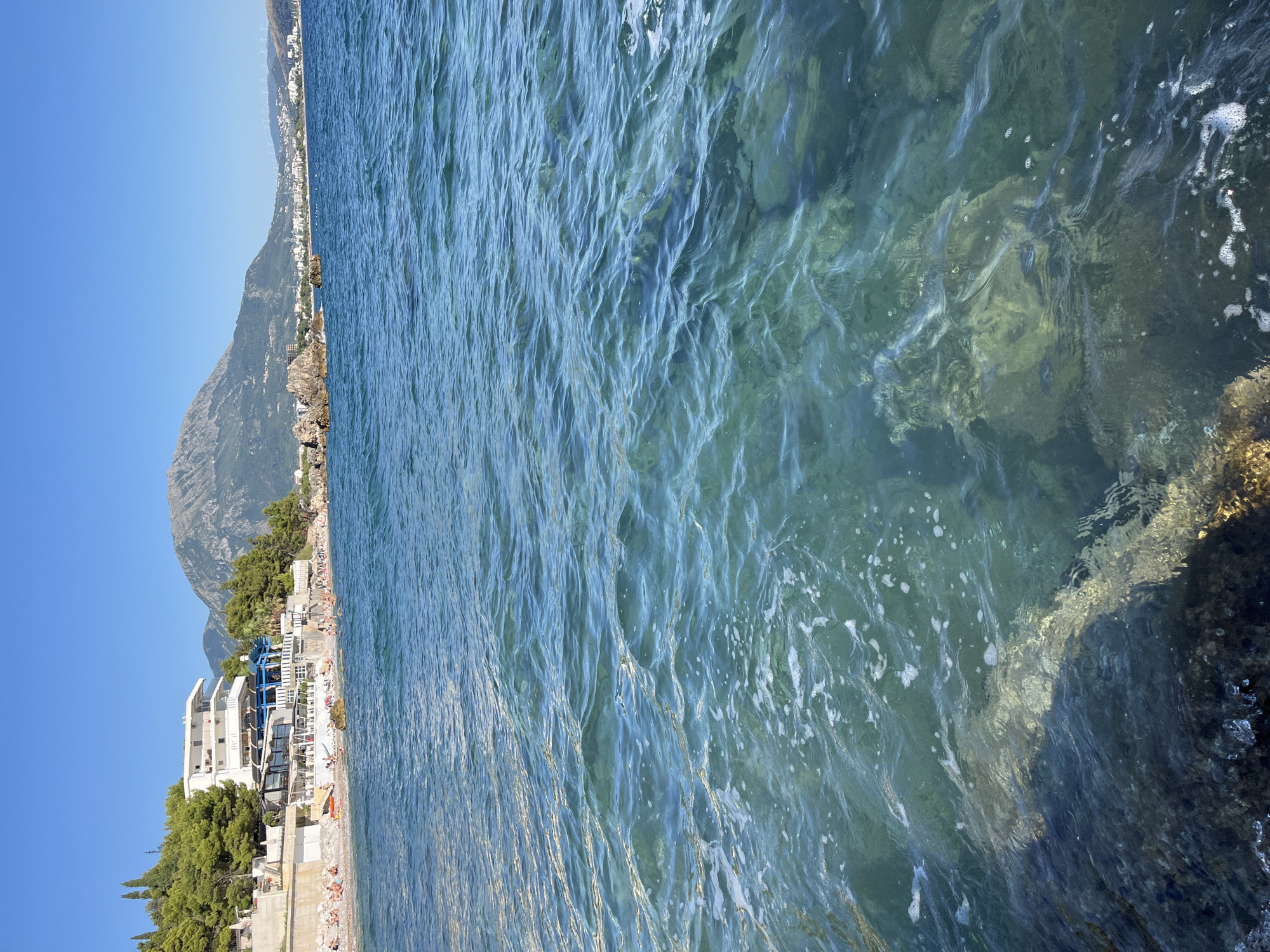 The image depicts a scenic view of the Adriatic Sea in Bar, Montenegro, with the shoreline and a town in the distance. In the foreground, the clear turquoise water is slightly rippled, revealing submerged rocks and pebbles. The shoreline is lined with a beach populated by numerous people sunbathing and swimming. A white building with blue accents, likely a hotel or restaurant, is situated on the elevated ground behind the beach, surrounded by lush green trees. Further in the background, a majestic mountain range rises, with the town of Bar sprawling along the coast. The sky is a clear, bright blue, indicating a sunny day.
