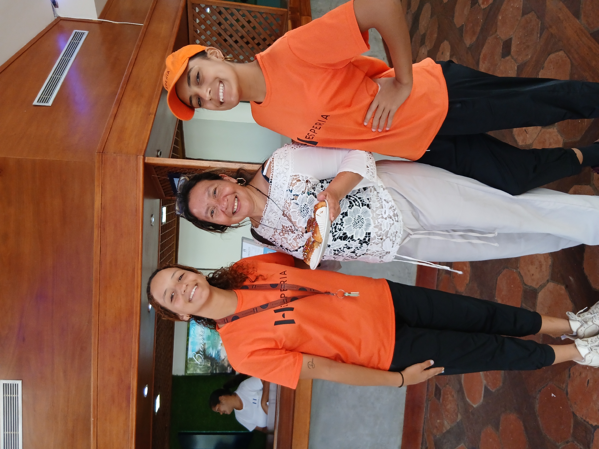 Three women are posing for a photo in an indoor space in Playa El Agua, Venezuela. The two women on the top and bottom are wearing matching bright orange shirts, black pants, and white shoes. The woman on top is also wearing an orange baseball cap. The woman in the middle is wearing a white lace top and white pants, and holding a plate with food. They are all smiling. The space has a wooden structure visible in the background, including a latticed section. The floor is tiled with red and brown geometric shapes. A person in a white shirt is visible in the background through a window.