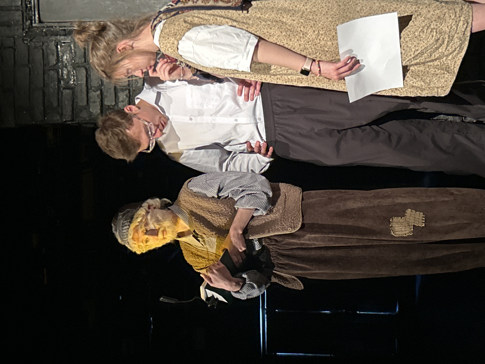 The image, captured in Unknown, Unknown, depicts three individuals on a dimly lit stage or performance area. On the left, an individual costumed as an older person, featuring a light-colored wig, beard, and facial makeup, wears a grey knit beanie, a blue and white plaid shirt, a brown fleece vest, and brown corduroy trousers with a light-colored, textured patch on the left thigh. This person holds an open, dark-covered book in both hands, illuminated by stage lighting. A black microphone on a stand is positioned to their left. In the center stands a young adult male with short brown hair and clear-rimmed glasses, dressed in a white long-sleeve button-down shirt and dark grey trousers, with his hands on his hips and gaze directed downwards. On the right, a young adult female with light brown hair tied in a bun wears a light-colored, patterned dress over a white short-sleeve top, a light-colored smartwatch, and multiple beaded bracelets on her left wrist. She holds a blank white piece of paper in her right hand and has her left index finger near her chin, looking towards the individual on the left. The background is largely dark, with a dark painted brick wall visible on the right side of the frame.