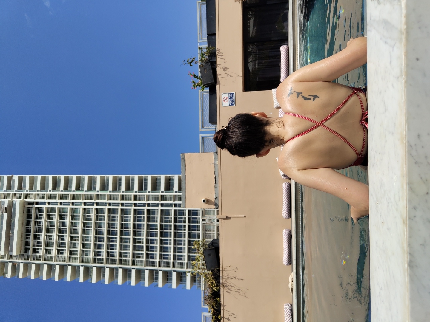 The image, oriented correctly, captures a sunny day scene at a rooftop pool overlooking an urban environment. In the foreground, a person is seen from the back, waist-deep in a swimming pool. The individual has dark hair pulled into a bun and is wearing a red and white striped bikini top with criss-cross straps. A tattoo depicting three stylized bird-like figures is visible on their upper back. Their skin appears wet and glistens in the sunlight. The edge of the pool in the immediate foreground is made of white marble.
Beyond the pool, a light brown building structure forms the base for the pool area. Along this structure, next to the water, are several rolled-up towels or cushions with a patterned design. To the far left of the image, a towering, modern high-rise building dominates the skyline, featuring numerous windows and balconies. The background is filled with a vast, clear blue sky, indicating bright, fair weather. Some greenery and potted plants are visible on the brown building structure.
A white sign with red iconography, likely a safety warning such as "no diving," is partially visible on the brown wall next to the pool. The scene suggests a relaxing day by the pool in a city setting, specifically Tel-Aviv, Israel, as per the provided context. The time of day is clearly daytime, with strong sunlight creating clear shadows. No specific emotions are discernible from the person's back view.