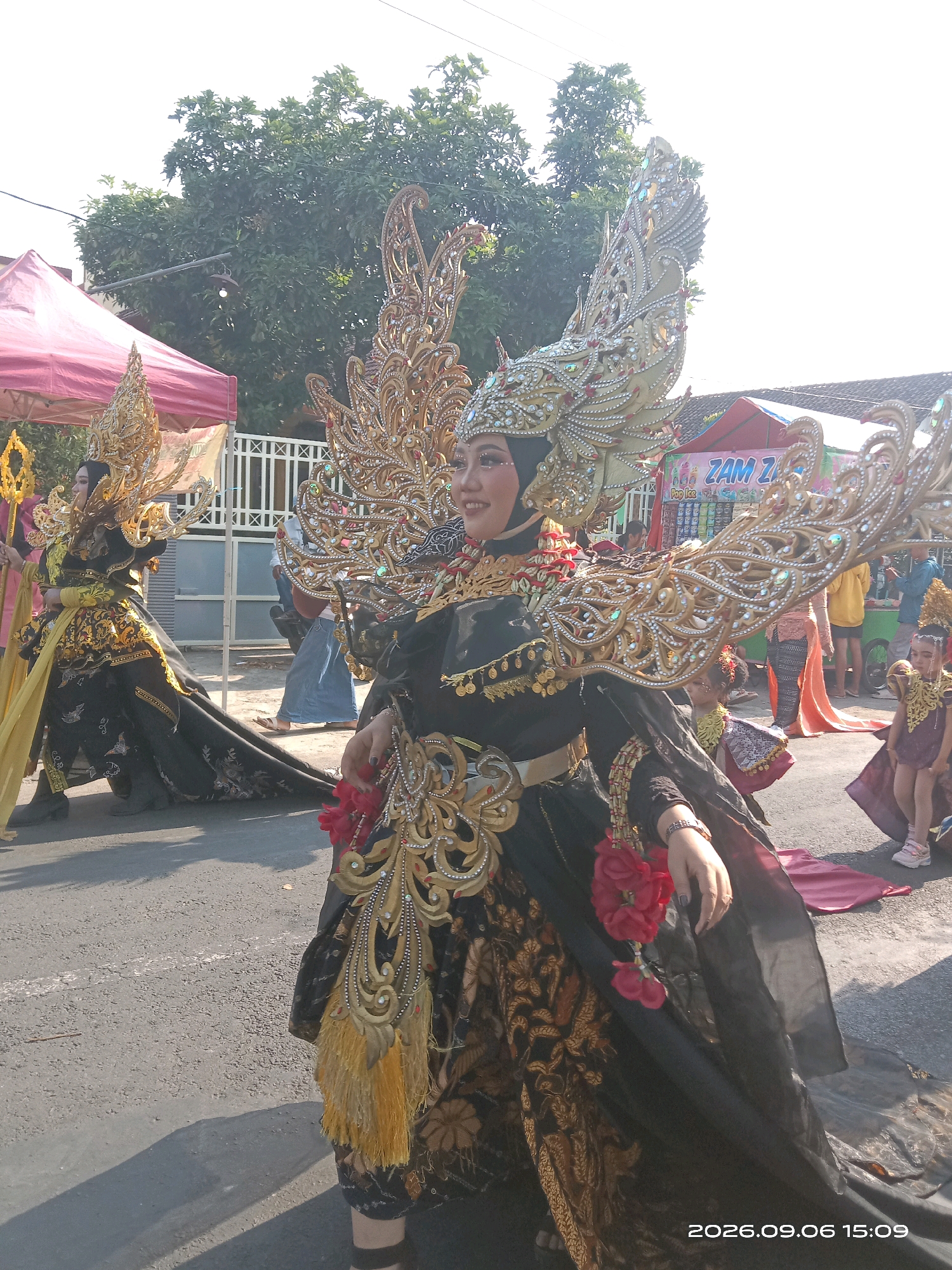 This is a medium shot, eye-level, outdoor, daytime photograph of a parade in Sepanyul, Indonesia. The foreground is dominated by a woman in elaborate, gold and black traditional Indonesian attire. She wears a large, ornate headdress with gold leaf detailing and colorful jewels. Her dress is black with gold patterns and is adorned with gold embellishments, red flowers, and tassels. She is smiling and looking to her left, with her right hand reaching out. To her left, another woman in a similar, but less ornate costume, is also wearing a gold headdress and a yellow sash. She is holding a golden staff. In the background, there are several other people, including children in traditional clothing. There are also tents and what appears to be a food stall with the sign "ZAM ZAM". The ground is asphalt, and there are shadows cast by the people and tents. The sky is bright and sunny. The date and time stamp on the image reads "2026.09.06 15:09".