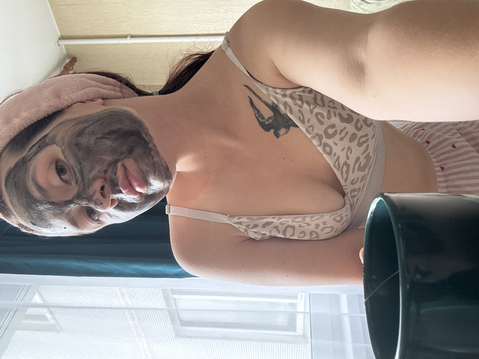 A young woman with a black face mask applied to the left side of her face is holding a dark green mug. She is wearing a light-colored bra with a leopard print and pink and white striped pajama bottoms. A pink towel is wrapped around her head. A tattoo of a bird is visible on her chest. She is indoors, in front of a window with sheer curtains. The location is described as Naberezhnye Chelny, Russia.