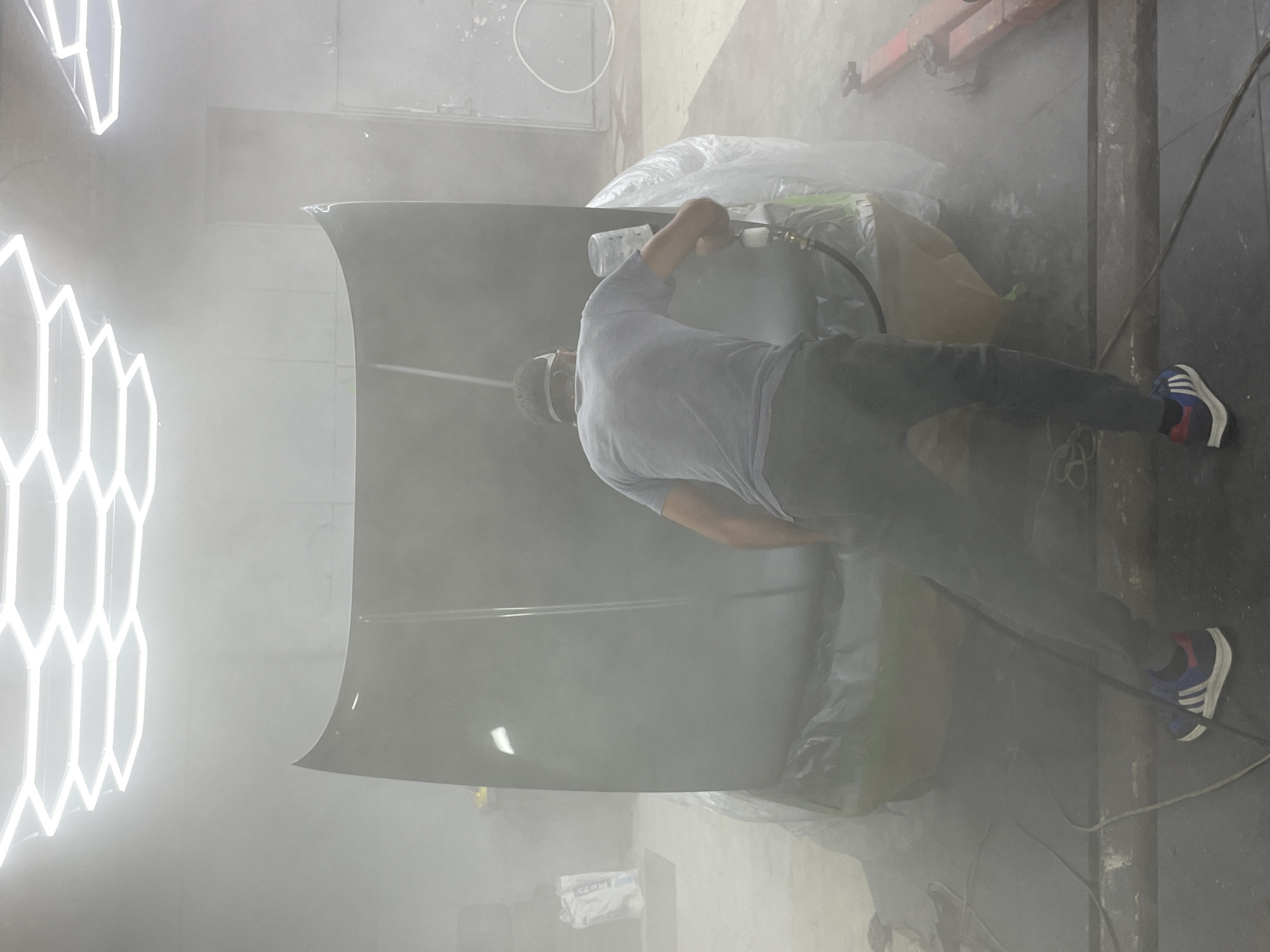 The image shows a person spray painting a car hood in a workshop. The person is wearing a grey t-shirt, dark pants, protective eyewear, and sneakers. They are holding a spray gun and appear to be in the process of applying paint to the hood, which is resting on sawhorses and covered in plastic sheeting. The workshop is filled with a mist of paint, creating a hazy atmosphere.
In the background, there are industrial elements such as a red overhead crane and various tools and equipment. The lighting in the workshop is a mix of artificial lights, including bright, geometric neon fixtures on the left side of the image. The overall impression is that of a busy auto body shop or a custom car painting facility. The presence of protective coverings and the spray of paint suggest that a significant amount of work is being done on the vehicle. There is no visible text in the image.