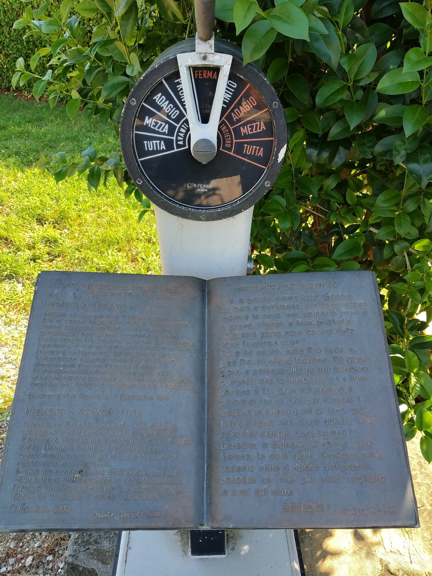The image depicts an outdoor memorial with a plaque resembling an open book and a ship's engine telegraph. The memorial is situated in front of lush green foliage and a patch of sunlit grass.
The engine telegraph, positioned above the plaque, is a circular dial with a lever. The dial is black with white and orange markings, including "FERMA" (STOP), "ADAGIO" (SLOW), "MEZZA" (HALF), and "TUTTA" (FULL) repeated for forward and reverse directions. The brand "R.FERRO GENOVA" is visible at the bottom of the dial.
The plaque, made of dark metal, is shaped like an open book. It contains inscriptions in Italian. The left side of the plaque features text with a heading "MEDAGLIA D'ARGENTO al V.M." followed by biographical details and a commendation for Giovanni Bizza, born in 1915 and deceased in 1992. The right side of the plaque bears the heading "PREGHIERA DEL MARINAIO" (Sailor's Prayer) and is followed by a prayer in Italian. Further down on the left side of the plaque, there is a heading "MEDAGLIA di BRONZO al V.M." and a commendation for an unnamed officer, dated September 1943. The bottom of the plaque shows "Orio Carrold 2003" and "Orio Carrold 2003" at the very bottom right. The scene appears to be a memorial site.