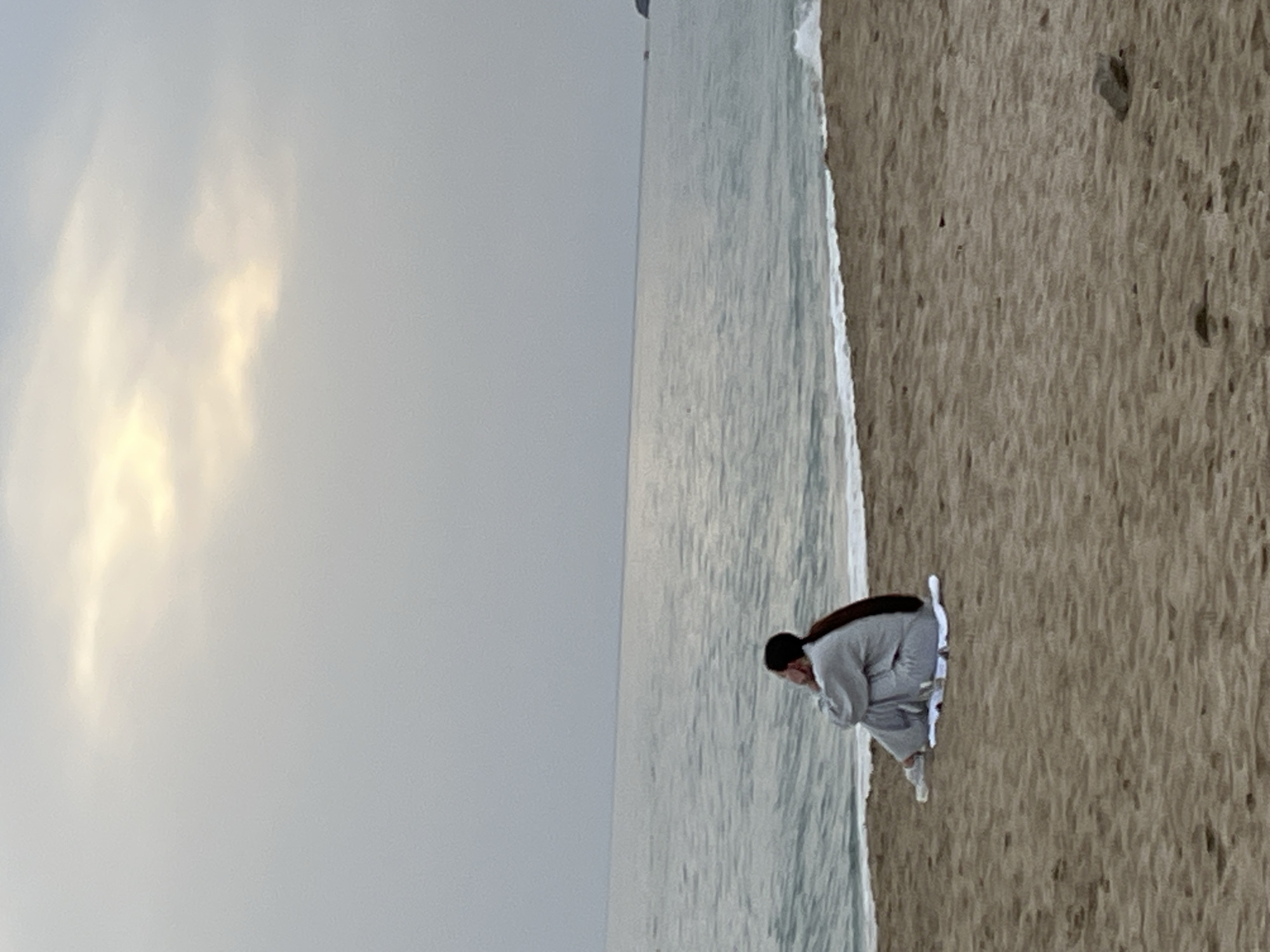 This photograph, taken in Alanya, Turkey, captures a serene coastal scene under an overcast sky. The horizon line neatly divides the muted grey sky, which features faint patches of diffused light from obscured clouds, from the calm, grey-blue sea. A thin line of white foamy waves gently laps at the light brown sandy shore.
In the foreground, positioned on the sandy beach near the water's edge, a single individual is visible. The person, appearing to be female with dark hair tied back, is dressed in a light grey long-sleeved top and matching long pants, possibly a casual tracksuit, with white sneakers. She is captured in a low crouching or kneeling posture, looking intently downwards towards the sand or the shallow water. Her posture suggests a moment of quiet contemplation, observation, or perhaps searching for something. The diffused light indicates it is daytime, though not bright and sunny. There is no visible text in the image. The overall impression is one of peaceful solitude on the beach.
