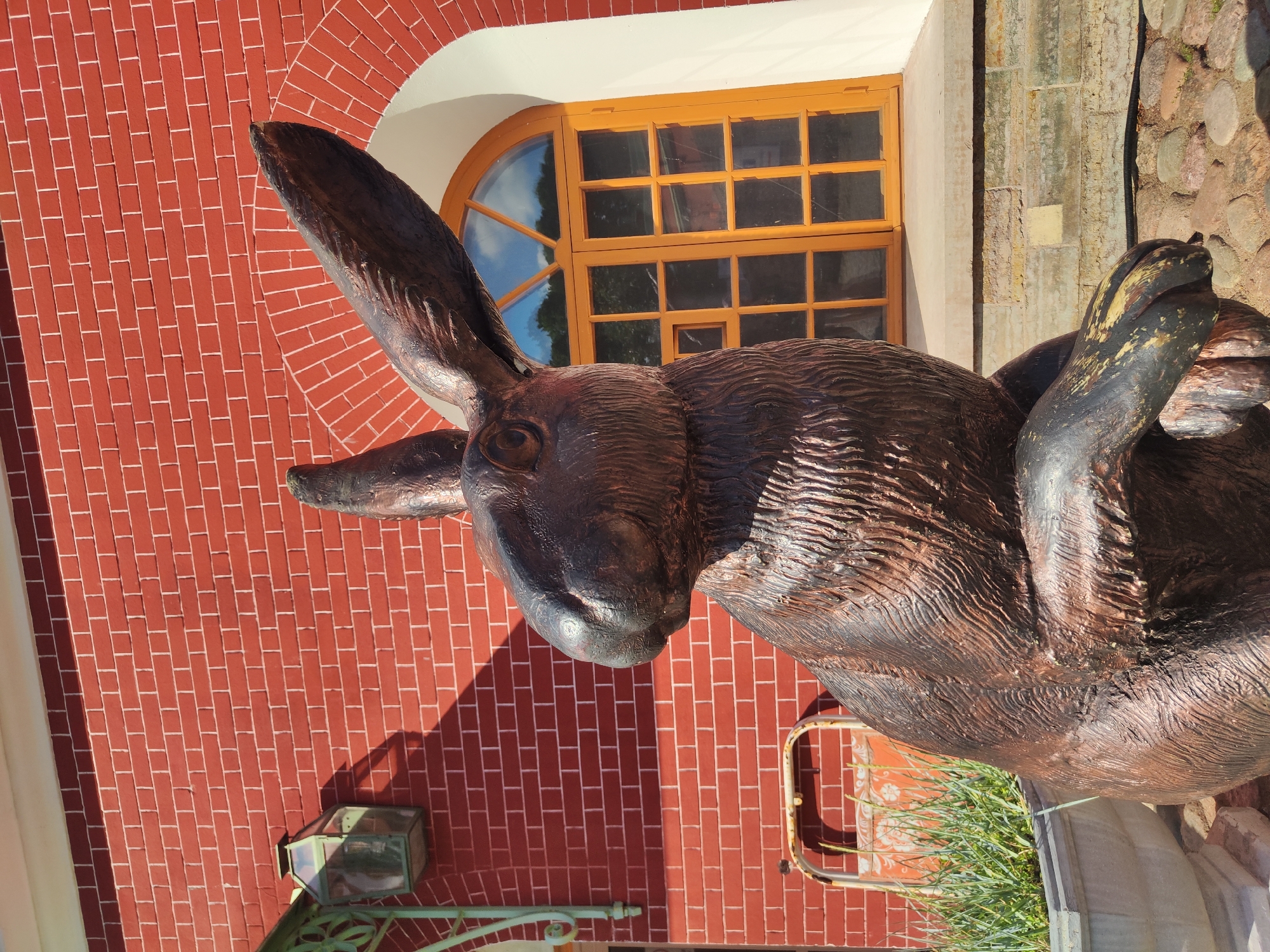 An outdoor, eye-level shot captures a bronze statue of a rabbit in front of a brick building. The rabbit is positioned in the foreground, facing left with its ears erect. The building behind it features a red brick facade, a dark green lantern mounted on the left side, and an arched window with an orange frame filled with glass panes. The window reflects a blue sky with some clouds and greenery. The base of the building is a light-colored stone ledge, and the ground in front of it is covered with small, rounded stones. To the left of the rabbit, a large, weathered planter holds green foliage. A rusted metal object with a floral pattern is visible behind the planter. The lighting suggests a sunny day, with shadows cast on the brick wall by the lantern and the rabbit statue. The scene appears to be in a park or garden setting.