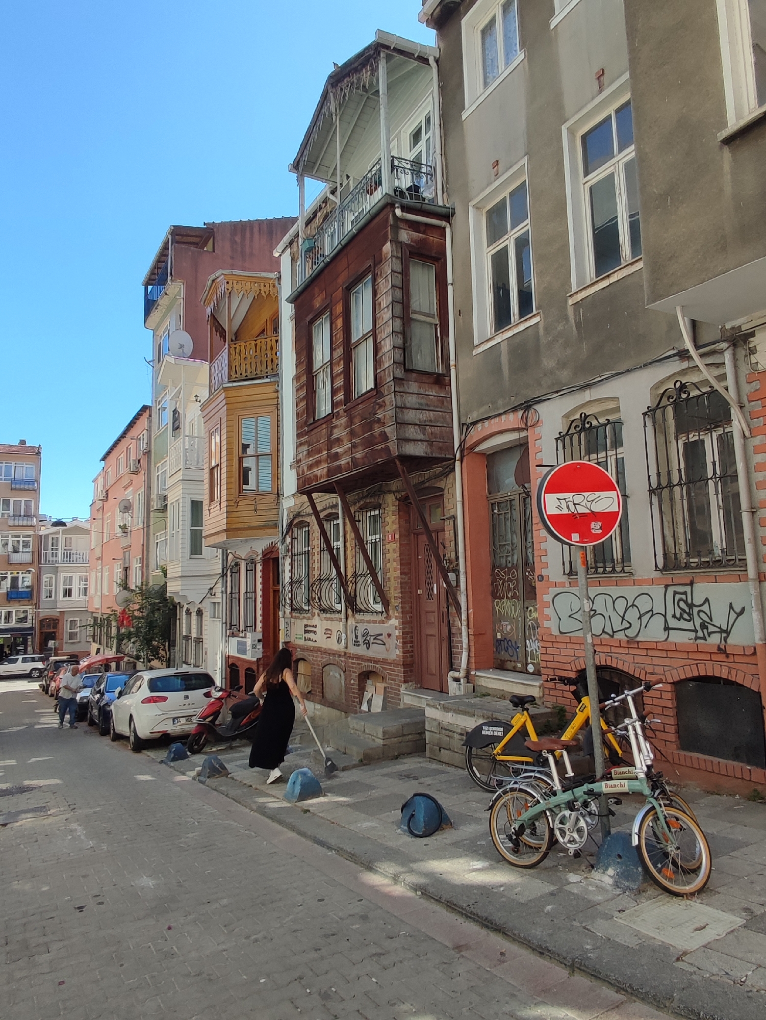 The image depicts a street scene in Kadıköy, Turkey. In the foreground, a woman dressed in a long black dress and white sneakers is sweeping the sidewalk with a broom. Two bicycles are parked on the sidewalk next to her. One is a yellow commuter bike, and the other is a light green foldable bicycle with "Bianchi" branding. A red and white circular traffic sign is mounted on a pole nearby. The background features a row of buildings with colorful facades and traditional architectural elements, including wooden balconies and bay windows. Several cars and a motorcycle are parked along the street. The sky is clear and blue.