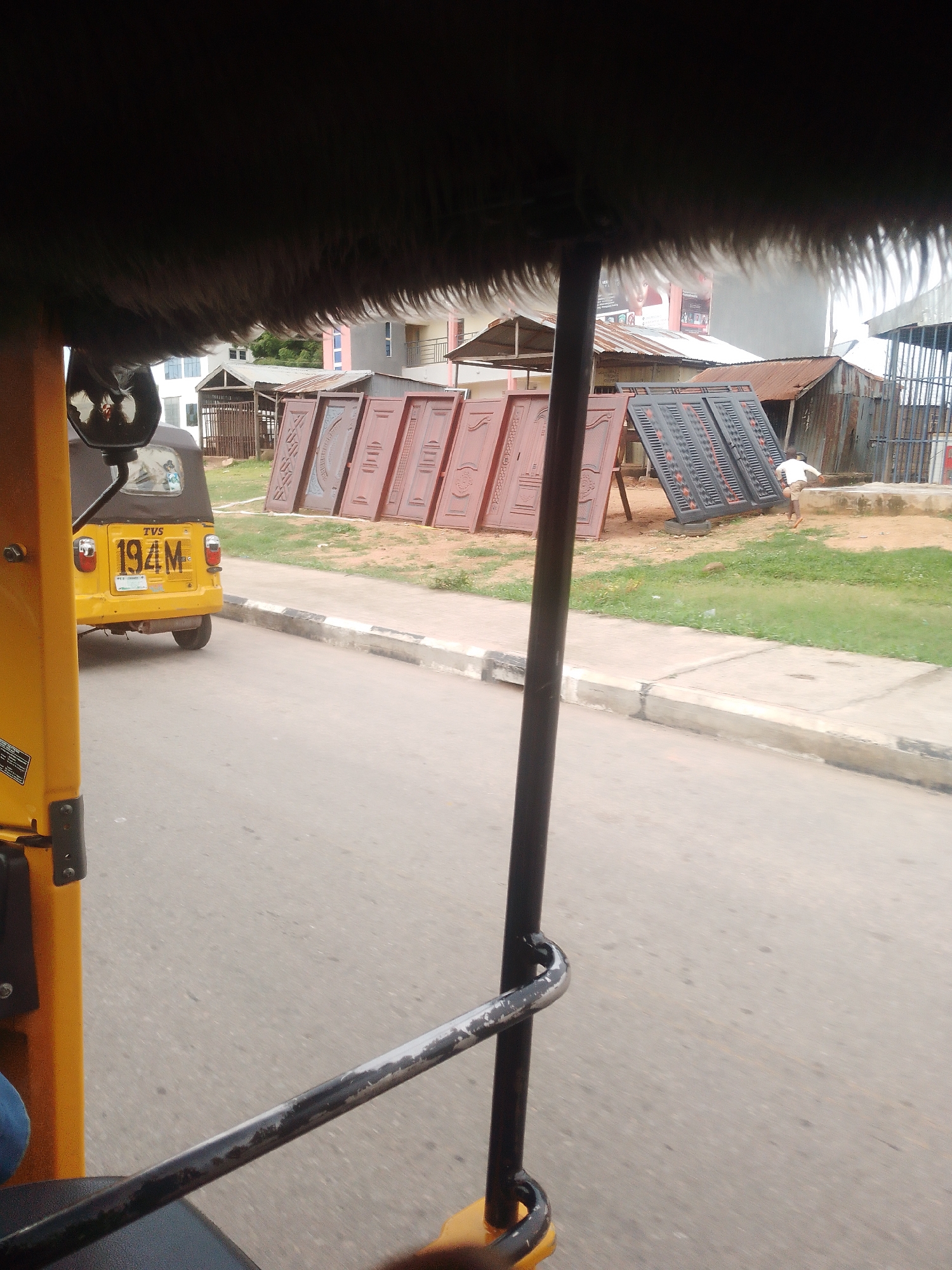 A low-angle, exterior shot captured from inside a yellow auto-rickshaw shows a street scene with several wooden and metal doors displayed for sale. A single person, a young male wearing a white t-shirt and brown shorts, is seen in the background interacting with the doors. The auto-rickshaw has a license plate that reads "194M". The environment includes a paved road, a sidewalk, a grassy area, and several buildings in the background, including what appears to be a multi-story residential building. The lighting suggests it is daytime.