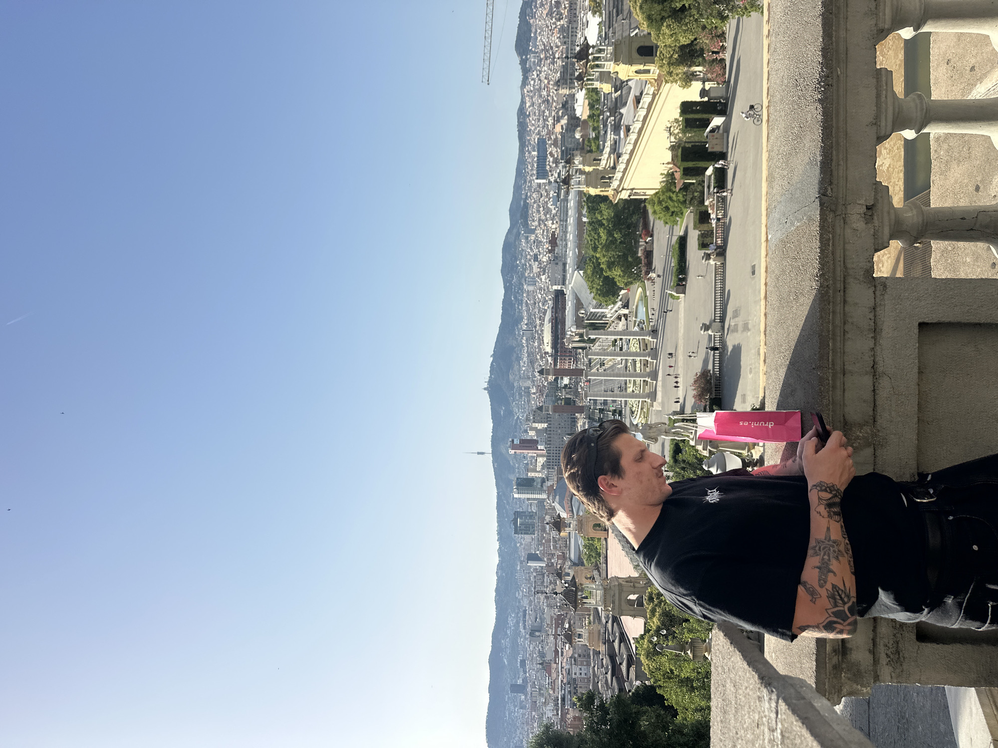 The image captures a man in a black t-shirt and black pants, with tattoos on his left arm, standing on a stone balcony overlooking a panoramic view of Barcelona, Spain. He is holding a pink water bottle and a smartphone, and appears to be looking at the phone. In the background, the cityscape unfolds with numerous buildings, a stadium, and a distant mountain range under a clear blue sky. The architectural style of the buildings suggests a European city, and the prominent structures in the middle ground, including colonnades and fountains, are indicative of a public square or monument area within Barcelona, possibly Montjuïc. The scene is bathed in sunlight, indicating daytime.