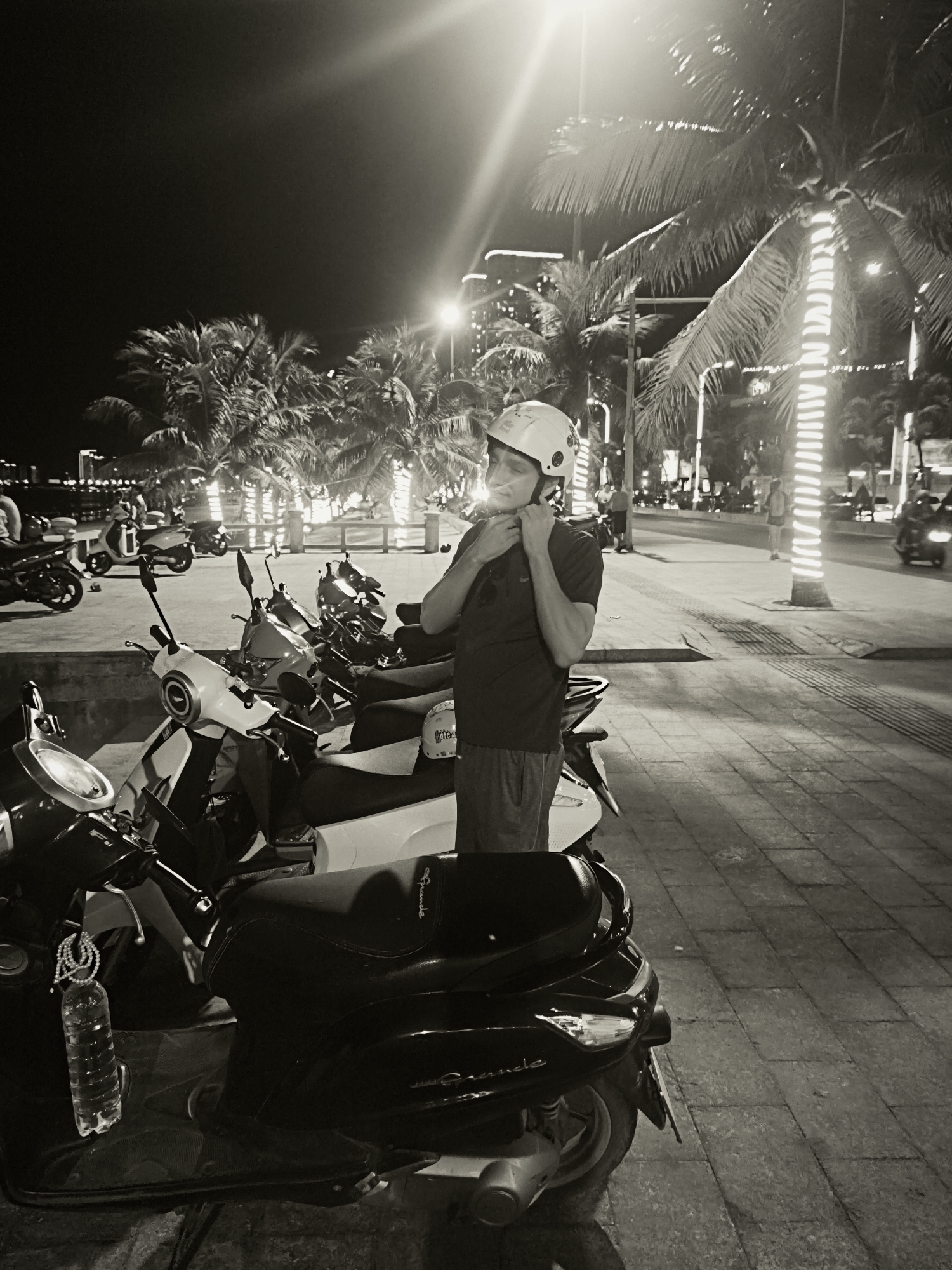 In a black and white nighttime scene on a paved sidewalk in Nha Trang Ward, Vietnam, a man stands adjusting a white helmet. He is wearing a dark t-shirt and shorts, and is positioned next to a row of parked scooters. Several palm trees are visible in the background, some adorned with string lights. A street with distant city lights and passing vehicles is also present. The atmosphere is illuminated by streetlights and the lights on the palm trees.