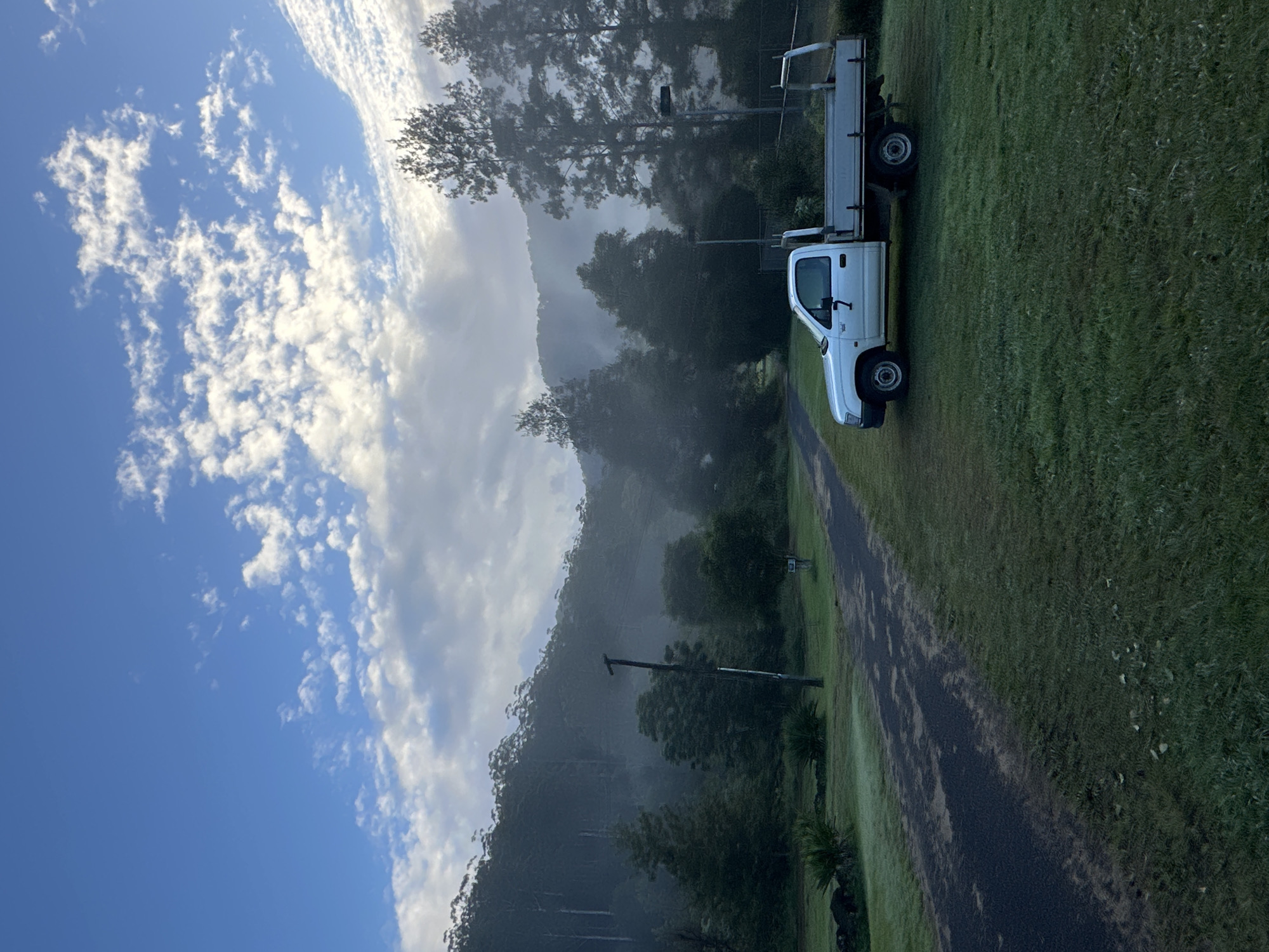 A white utility truck is parked on a grassy area to the right of a paved road. The road leads away from the viewer and curves to the left. The truck is facing to the right. In the background, there is a dense forest on a misty mountainside under a sky with scattered clouds. A wooden pole with what appears to be electrical wires attached stands near the left edge of the road. The scene is set in Thora, Australia.