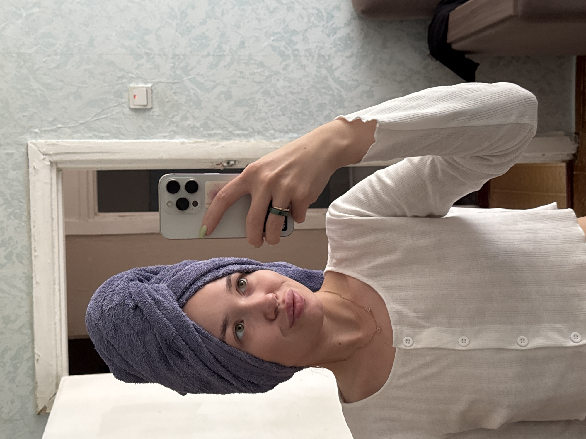 A young adult female is depicted in a mirror selfie, standing in a room in the city of Chaikovsky, Russia. She has her hair wrapped in a light purple, textured terry cloth towel, turban-style. Her lips are slightly puckered, and her green eyes gaze upward and to the right, past the camera. She wears a white, long-sleeved, ribbed or textured shirt with a rounded neckline and a vertical row of white buttons down the front; the lowest visible button is undone. A thin gold necklace with small pendants, including one resembling the letter 'G', adorns her neck. Her right hand, with short nails painted a pale lime green, holds a silver-colored smartphone with a multi-lens camera array vertically to take the photo. A dark green band ring with an adjacent thin gold band is visible on her left ring finger.
The background, reflected in the mirror, reveals an aged interior. To the left, a white, worn door frame with peeling paint is visible, leading into a darker room beyond. The wall on the right is covered with light blue patterned wallpaper, possibly with a subtle floral or abstract design, and exhibits faint vertical lines or creases. A white light switch with a small red indicator light is mounted on this wall. Further to the right, a section of brown upholstered furniture, possibly a sofa, is visible with a black piece of clothing draped over it. The lower right portion of the reflection shows a tiled floor, appearing light brown or terracotta in color.