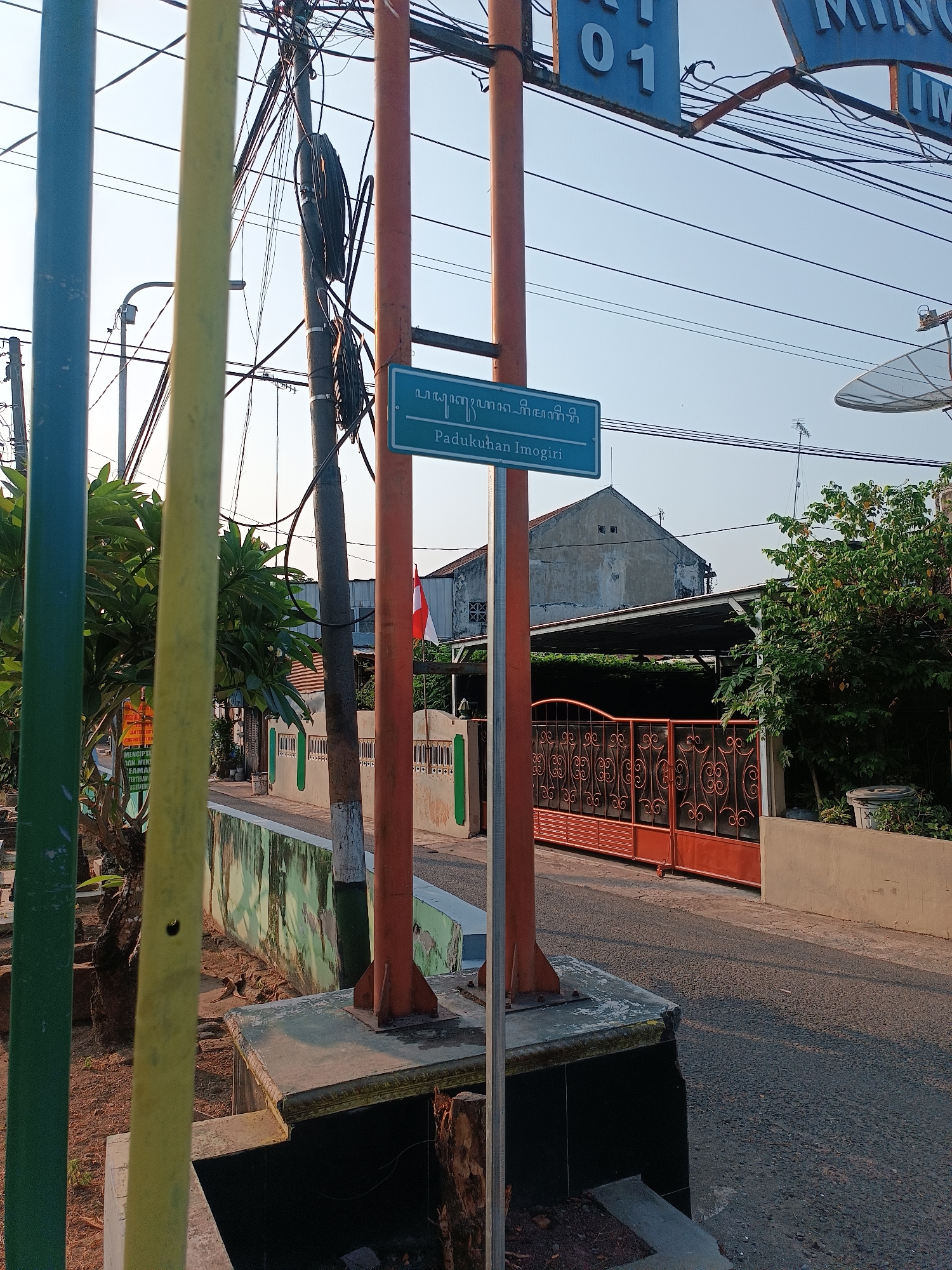 The image shows a street scene in Imogiri, Indonesia. In the foreground, there are several vertical poles, one colored green, one yellow, and two orange. Behind these poles, a blue street sign is visible, reading "Padukuhan Imogiri" in white text, with some script above it. The sign is mounted on a gray metal pole, which is next to one of the orange poles. Beyond the sign, a street runs past several houses and a bright orange gate with an ornate pattern. An Indonesian flag is visible on a flagpole in the background. The sky is clear and blue. Electrical wires and a satellite dish are also present in the scene. The overall impression is a quiet residential street in a tropical climate.