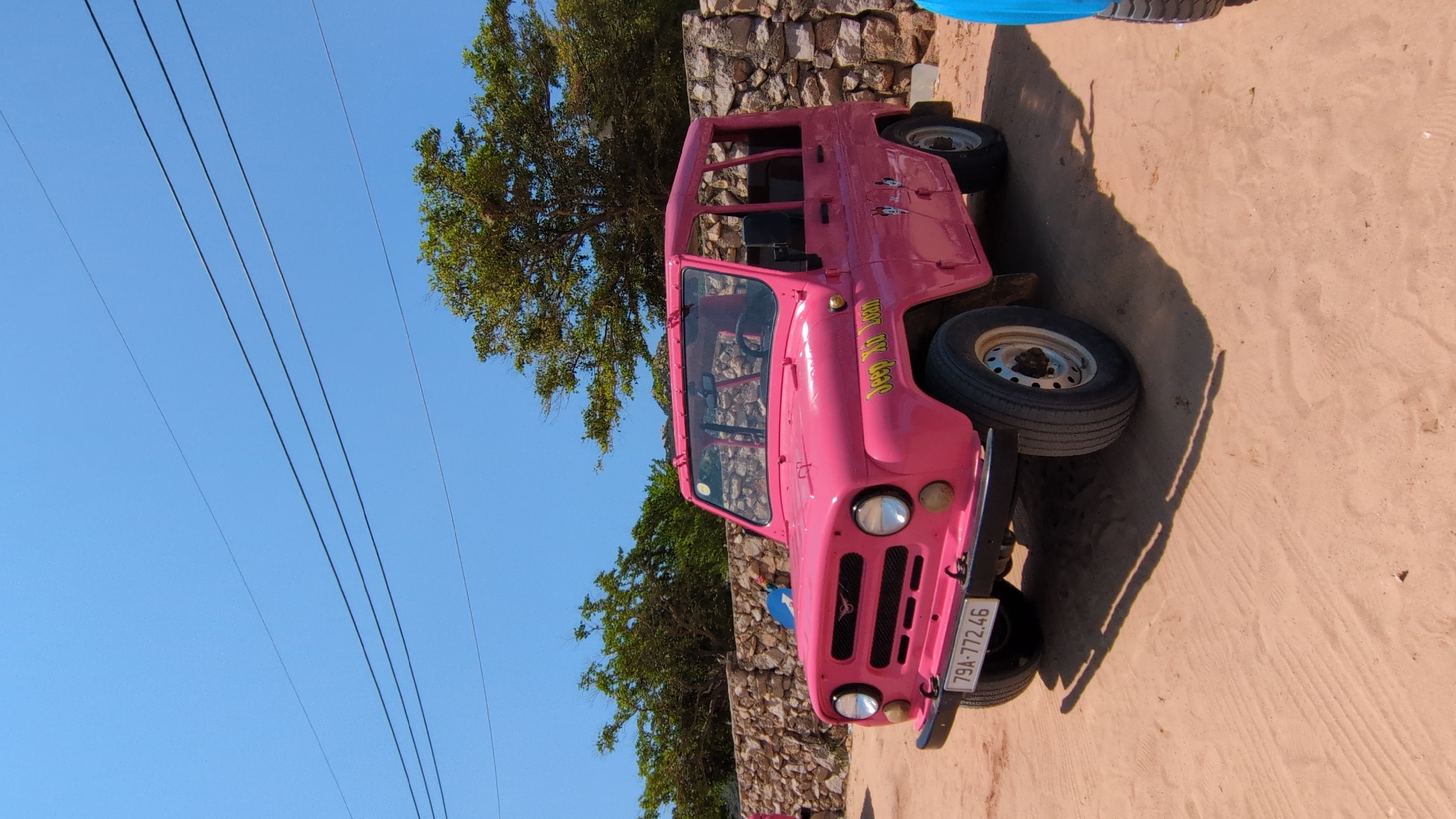 A pink UAZ-469 vehicle with license plate "79A-772.46" is parked near a stone wall in Phuoc Dinh, Vietnam. The vehicle is facing towards the camera and has a sign on the passenger side that reads "Jeep Air Tour". Power lines are visible against a clear blue sky. A tree grows beside the stone wall. A sandy ground is visible under the vehicle. A small portion of a blue object is visible near the top right corner of the image.