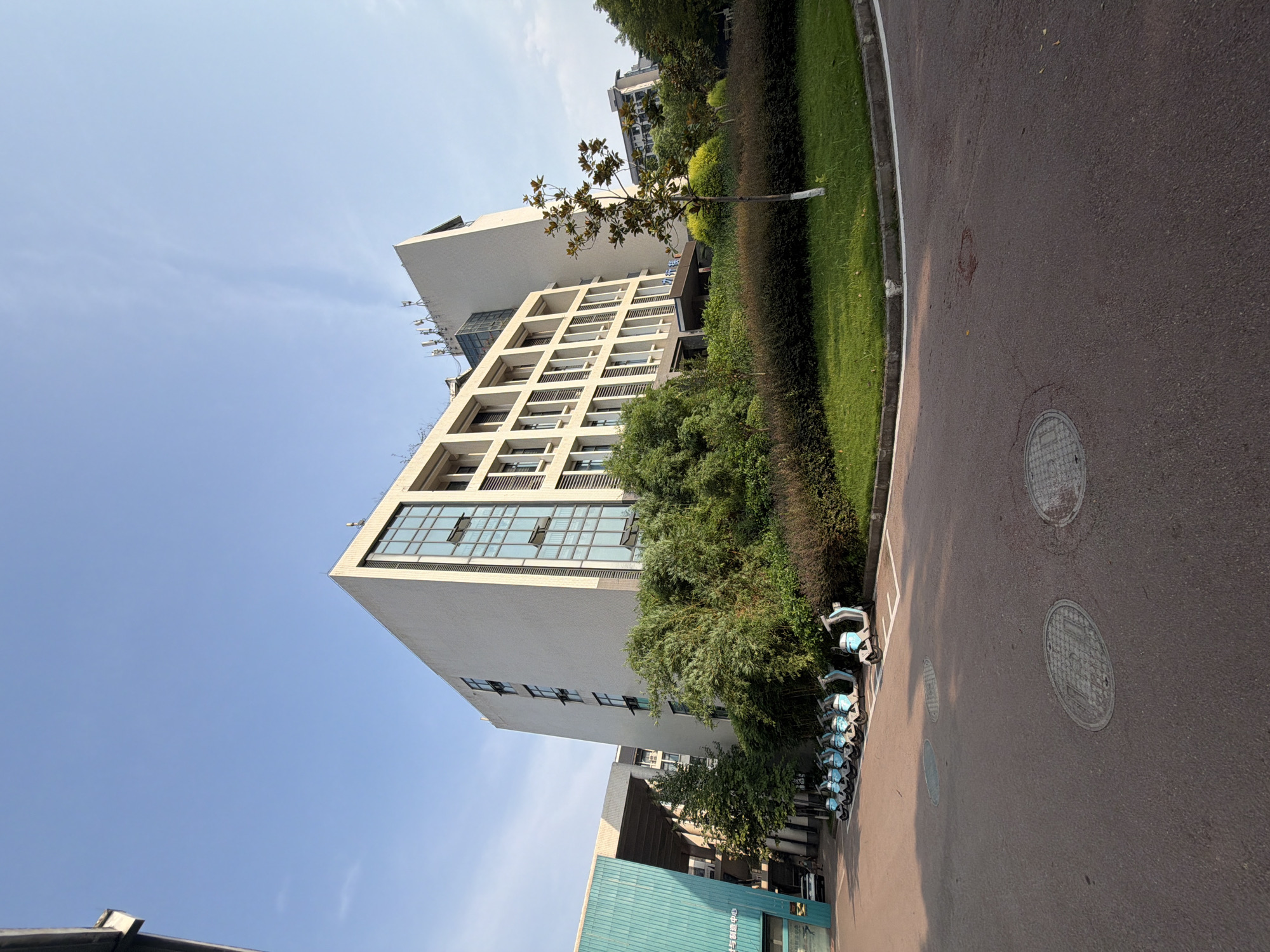 A low-angle, wide shot captures a modern, beige building with a prominent glass facade and a blue-green adjoining structure. A line of electric scooters is parked in front of the building. Lush green trees and manicured bushes surround the building, creating a park-like setting. The pavement in the foreground shows circular manhole covers. The sky is clear and blue, indicating a sunny day. The scene is located in Jiangjin, China. There are no people or animals visible in the image.
