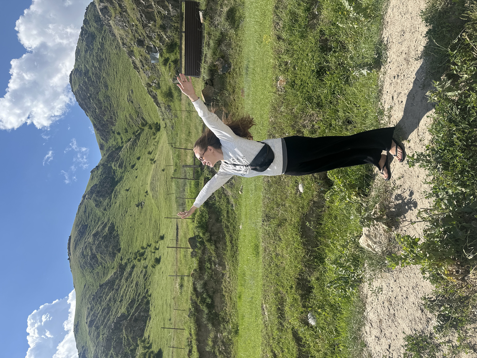 A young woman stands in a rural, mountainous landscape. She is facing away from the camera with her arms outstretched and head tilted back, appearing to embrace the scenery. She wears a grey sweatshirt, black pants, and black flip-flops. A black fanny pack is worn across her chest. Her long, brown hair is windblown. The background consists of rolling green hills and mountains under a bright blue sky with scattered clouds. A wire fence runs horizontally across the mid-ground. A dark brown, wooden structure is visible on the right side of the frame. The ground in the foreground is a mix of dirt and sparse vegetation. The location is identified as Bylym, Russia.