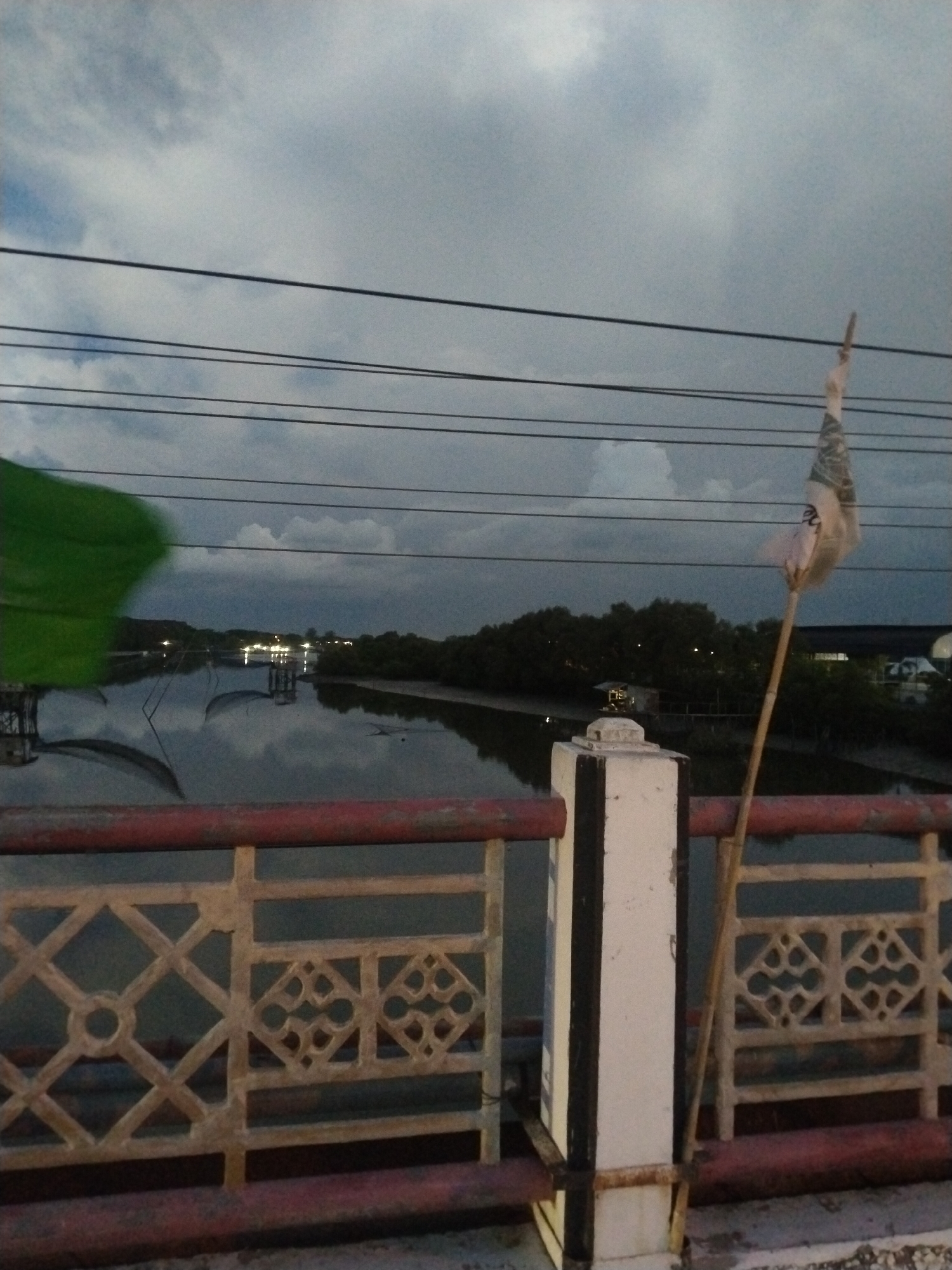 The image, likely taken in Lhokseumawe, Indonesia, shows a view from a bridge overlooking a body of water. The sky is overcast with dark clouds. Power lines stretch across the upper part of the frame. The bridge railing is painted red and white, with a decorative pattern of geometric shapes. A green cloth or flag is partially visible on the left side of the frame, moving in the wind. A tattered white flag is attached to a bamboo stick leaning against one of the bridge pillars on the right side. The water reflects the sky and the silhouettes of trees lining the distant shore. Lights are visible in the distance, suggesting a built-up area. A structure resembling a fishing platform is visible in the water.