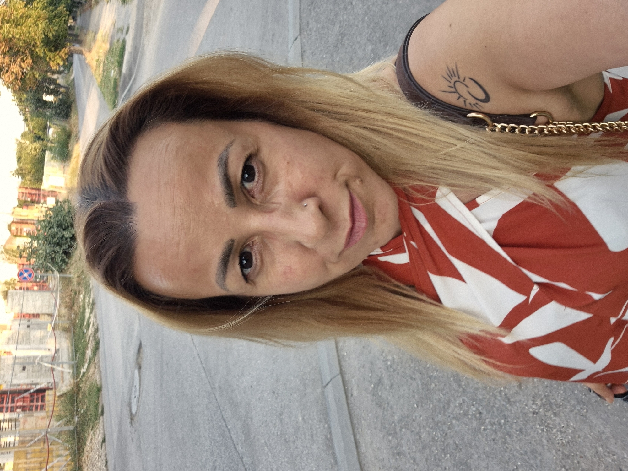 A woman with blonde hair and a nose ring is taking a selfie outdoors. She is wearing a red and white patterned top and has a tattoo on her right shoulder. The background appears to be a street scene with buildings and trees.