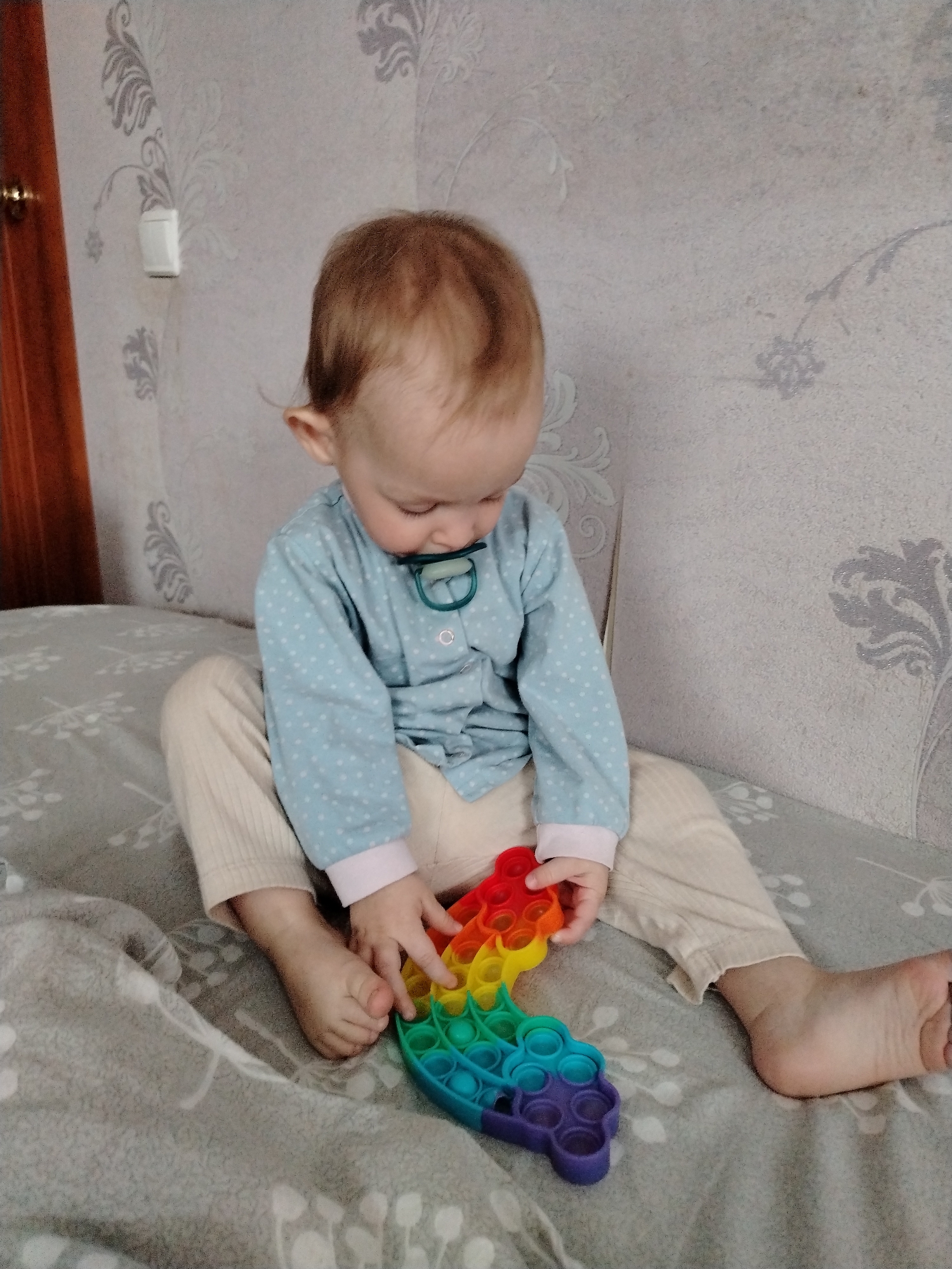 A young child is sitting on a bed in a room. The child has fair skin, light brown hair, and is wearing a light blue, long-sleeved shirt with white polka dots and light-colored pants. A pacifier is in the child's mouth. The child is focused on a rainbow-colored "pop-it" toy in their hands. The bed has a grey patterned bedspread. The walls are light-colored with a floral pattern. A wooden door frame is visible on the left. The image appears to be taken indoors with natural lighting. The location of Nizhnyaya Gar', Russia is not evident within the image.