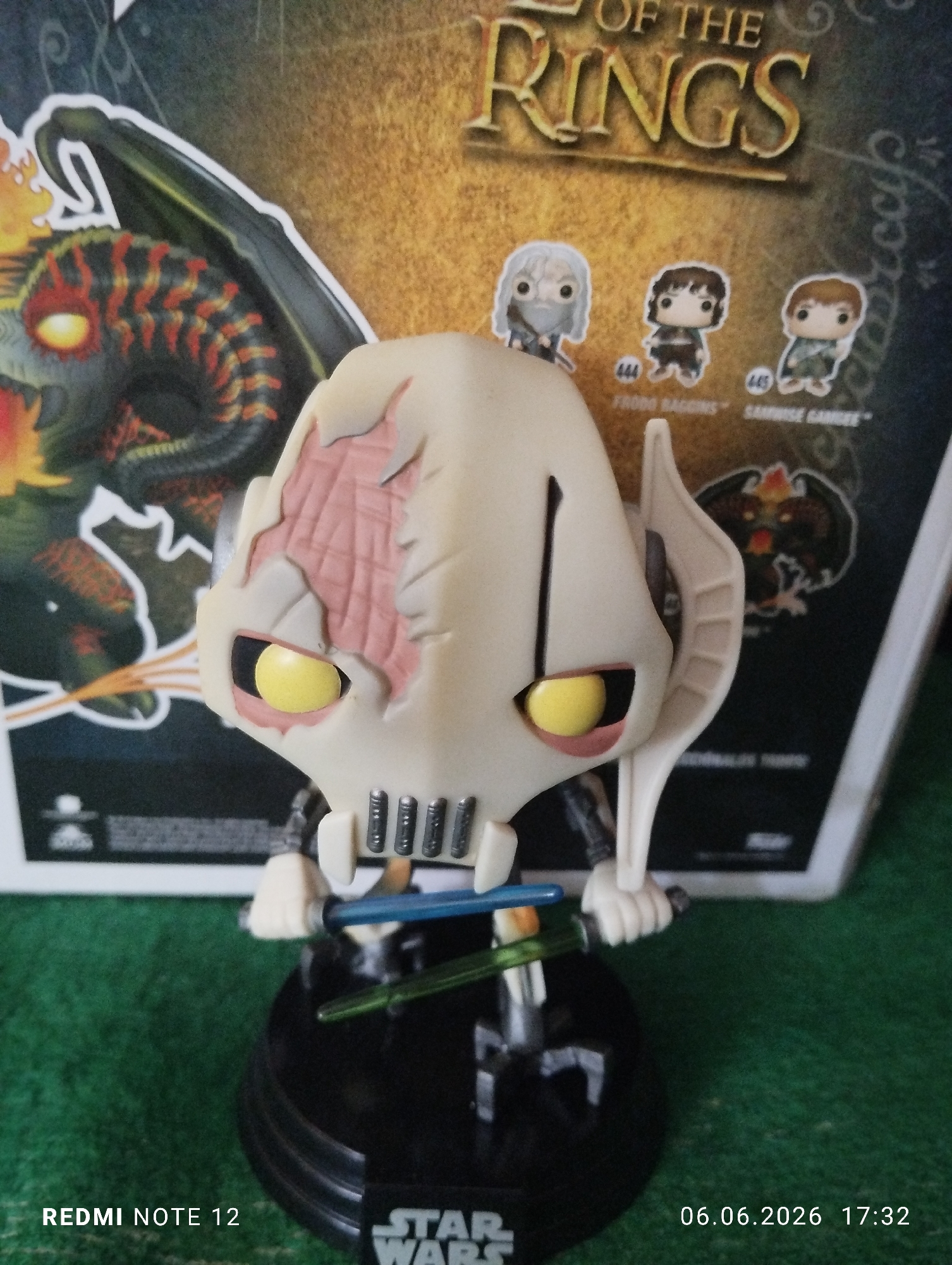 A close-up photograph, captured by a REDMI NOTE 12 device on June 6, 2026, at 17:32 in the city of Burgas, Bulgaria, displays a Funko Pop! vinyl figure of General Grievous from Star Wars. The figure is predominantly off-white and features four arms, each holding a lightsaber; one is blue, and the other is green. Its large head includes prominent yellow eyes and an exposed internal reddish-pink structure on the figure's proper right side. The General Grievous figure is positioned on a circular black base inscribed with "STAR WARS." In the background, partially out of focus, stands a cardboard box, identifiable by the text "OF THE RINGS," indicating a Lord of the Rings product. This box showcases an illustration of a dark, winged, dragon-like creature with glowing eyes on its left side, and three smaller Funko Pop! character images below the title: one masked figure, "444 FRODO BAGGINS," and "445 SAMWISE GAMGEE." The scene is set on a uniformly green, textured surface, resembling felt or short-pile carpet.