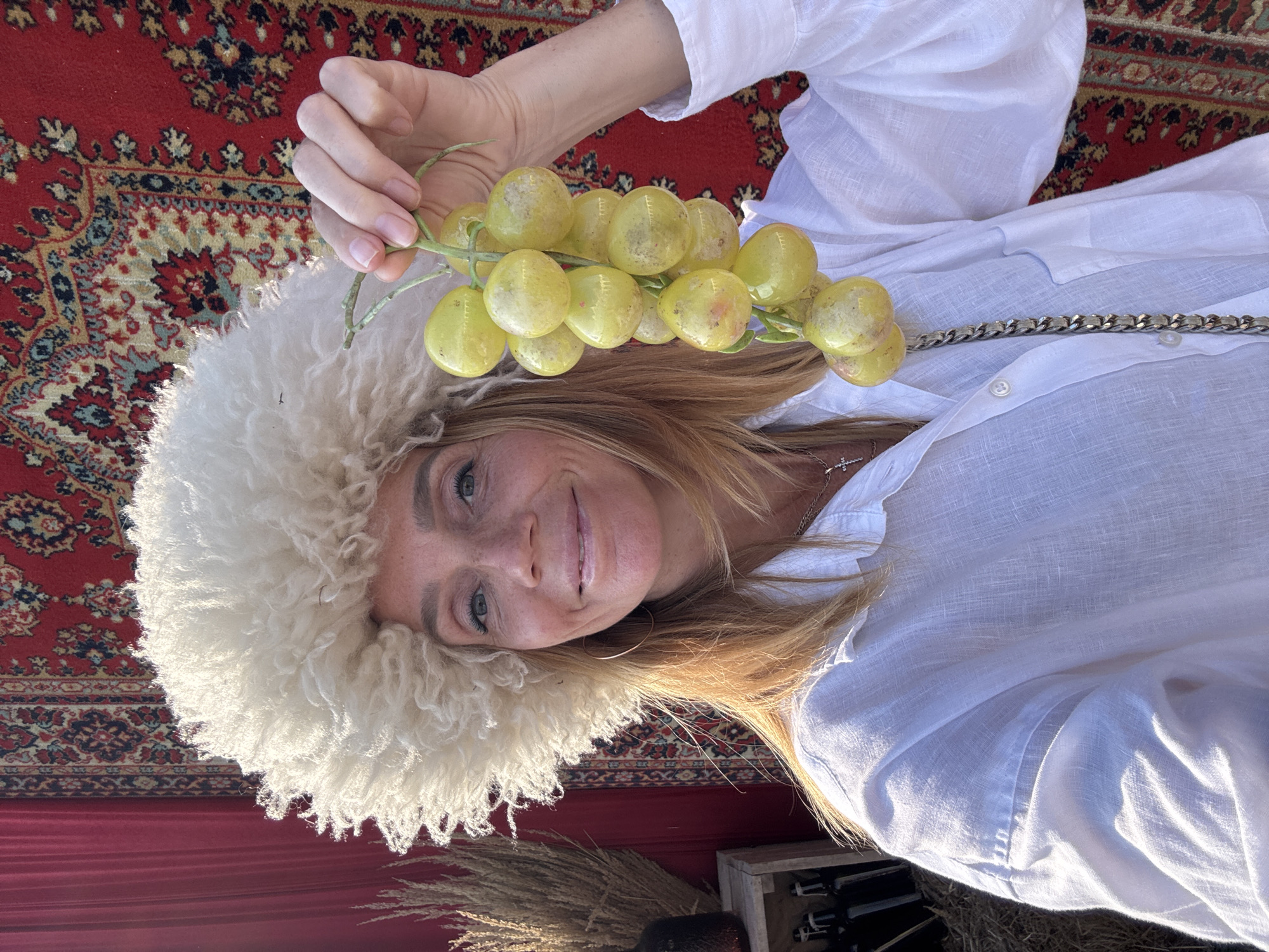A woman in a fluffy white hat and a white linen shirt holds a bunch of light green grapes. She is smiling and looking at the camera. The background features a red curtain and a patterned rug. The image appears to be a selfie taken outdoors during daylight. There are no discernible elements that indicate the location as Veliky Novgorod, Russia.