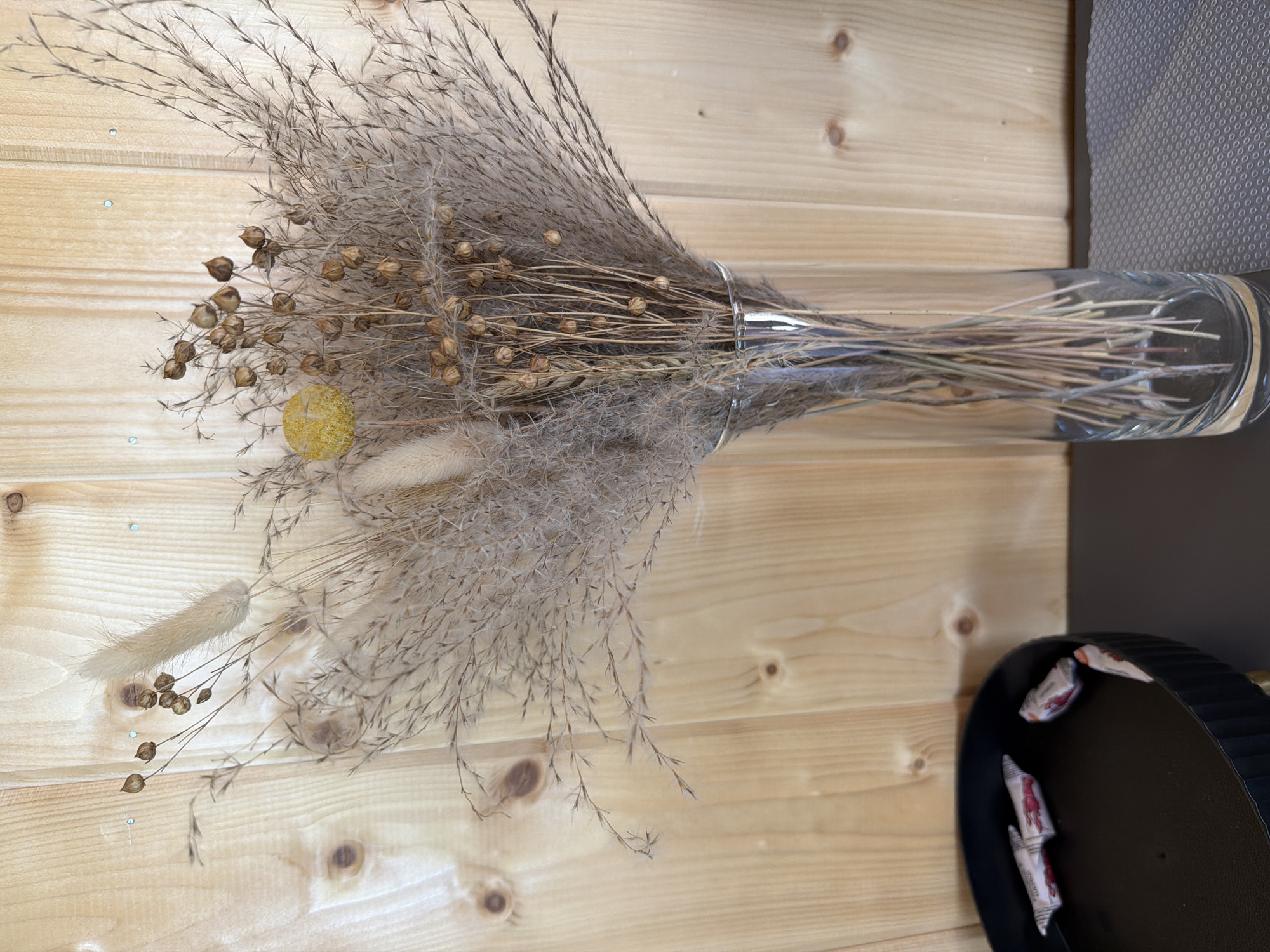 The image depicts a still life scene featuring a bouquet of dried flowers and grasses in a clear glass vase. The bouquet is composed of various textures and colors, including feathery plumes, thin stalks, and seed pods. A single yellow decorative sphere is also visible within the arrangement. The vase stands on a dark surface, possibly a table, to the right of a dark, ribbed bowl containing several individually wrapped candies. The background consists of a wooden paneled wall, exhibiting a light, natural wood grain. The lighting appears to be soft and diffused, casting subtle shadows. The overall impression is one of rustic, natural decoration. There is no indication of the city of Новый, Russia, or any specific urban environment within the image.