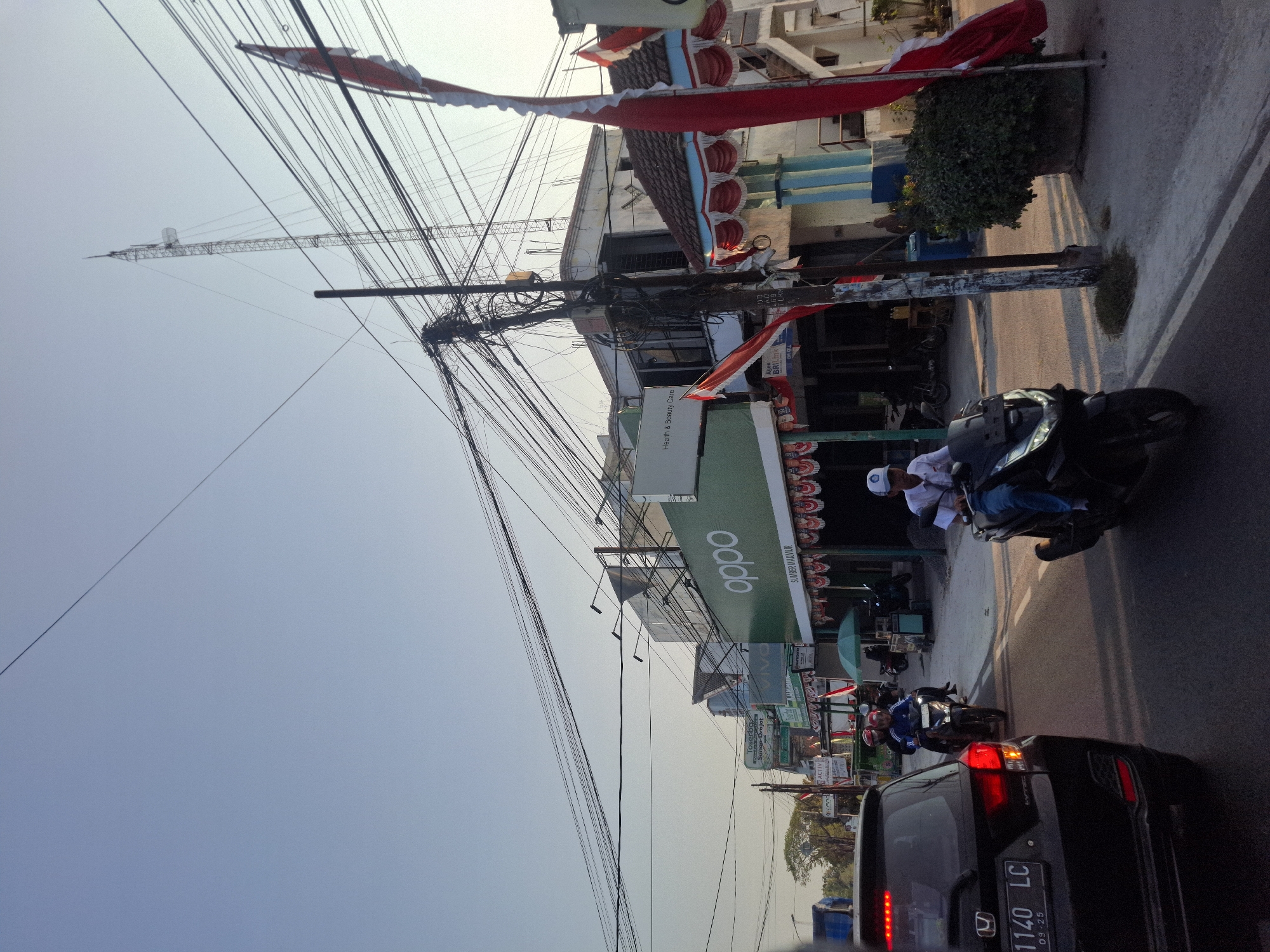 The image depicts a street scene in Mertani, Indonesia, likely during daylight hours, with clear skies. A black Honda car is stopped in traffic, its brake lights illuminated. In front of the car, several motorcycles are also in traffic, with riders wearing casual attire and helmets. The road is paved, and a sidewalk runs along the side of the street. Buildings with storefronts line the street, featuring prominent signage for "OPPO," "Toserba," and "Vivo." Indonesian flags, red and white, are displayed on poles and draped across some buildings, suggesting a national holiday or celebration. A tall telecommunications tower is visible in the background, surrounded by a dense network of electrical wires. Lush green trees are present on the left side of the image, contrasting with the urban environment.