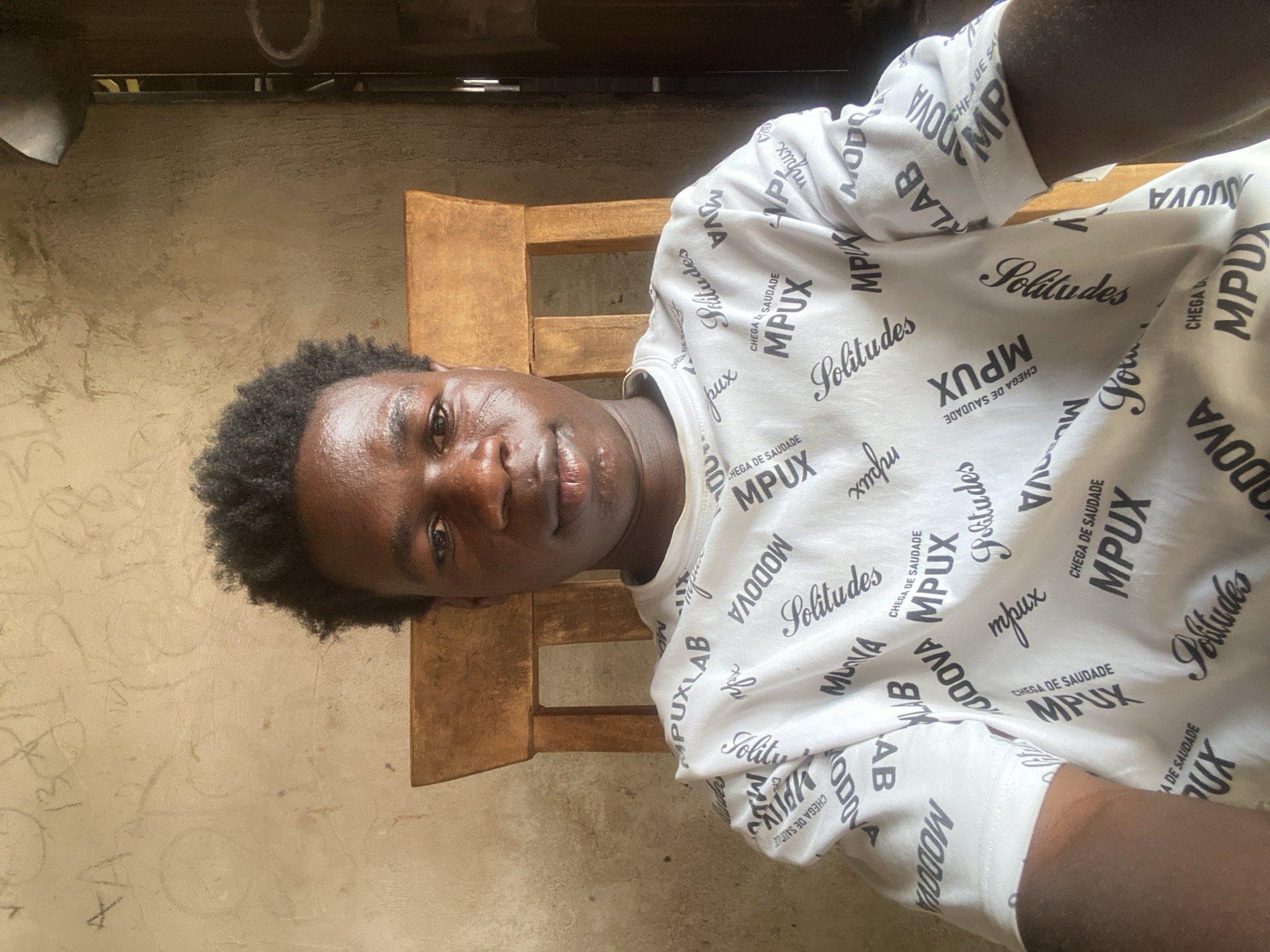 This media file presents a close-up portrait of a young Black man, taken in Mararaba, Nigeria. The man, positioned horizontally in the frame, looks directly at the camera with a neutral to slightly thoughtful expression. He has short, dark, curly hair and his skin appears well-hydrated.
He is wearing a white short-sleeved t-shirt adorned with a pattern of black text in various fonts and sizes. Prominent words and phrases repeated across the shirt include "Solitudes" (in a stylized script font), "MPUX" (in a bold, sans-serif font), and "CHEGA DE SAUDADE," which is Portuguese for "enough of longing" or "no more longing." Other visible text elements include "MODOVA," "MUVA," and fragments like "MPUXLAB" and "LAB."
The background consists of a light brown or beige textured wall, suggesting an indoor or semi-indoor setting. A portion of a wooden chair, likely its back or armrest, is visible behind him to the right. The lighting indicates it is daytime, with natural light illuminating the scene. The man appears to be posing for a photograph in a casual setting.