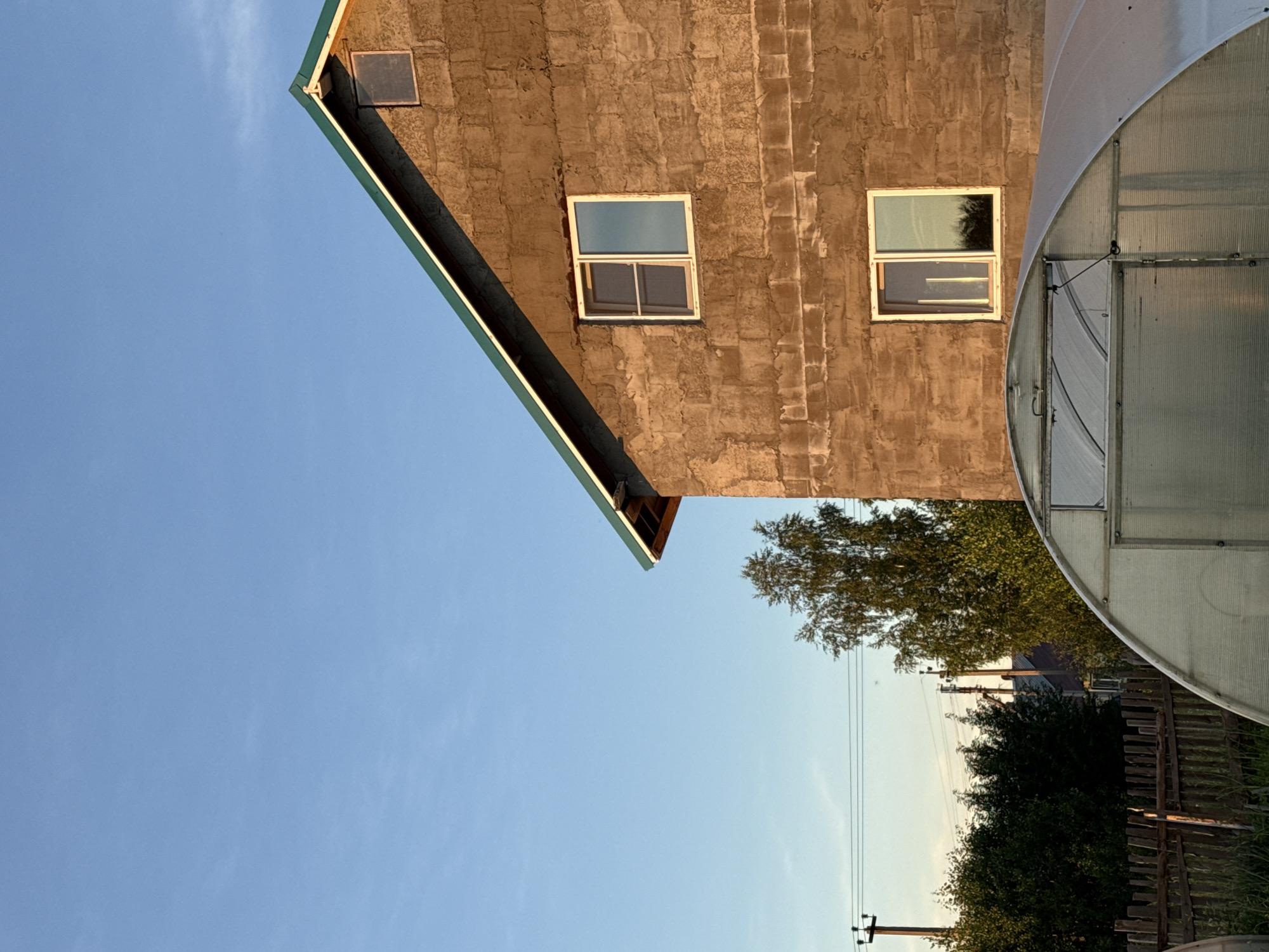 The image shows a two-story building with rough, brown walls, possibly under construction, with a green-sloped roof. There are at least three windows visible on the side of the building. In the foreground, there is a clear plastic greenhouse with a curved roof. Behind the building, there are trees, power lines, and a wooden fence. The sky is clear and blue. The scene appears to be in a rural or suburban area, likely in the village of Garevaya, Russia, given the architectural style and natural surroundings.