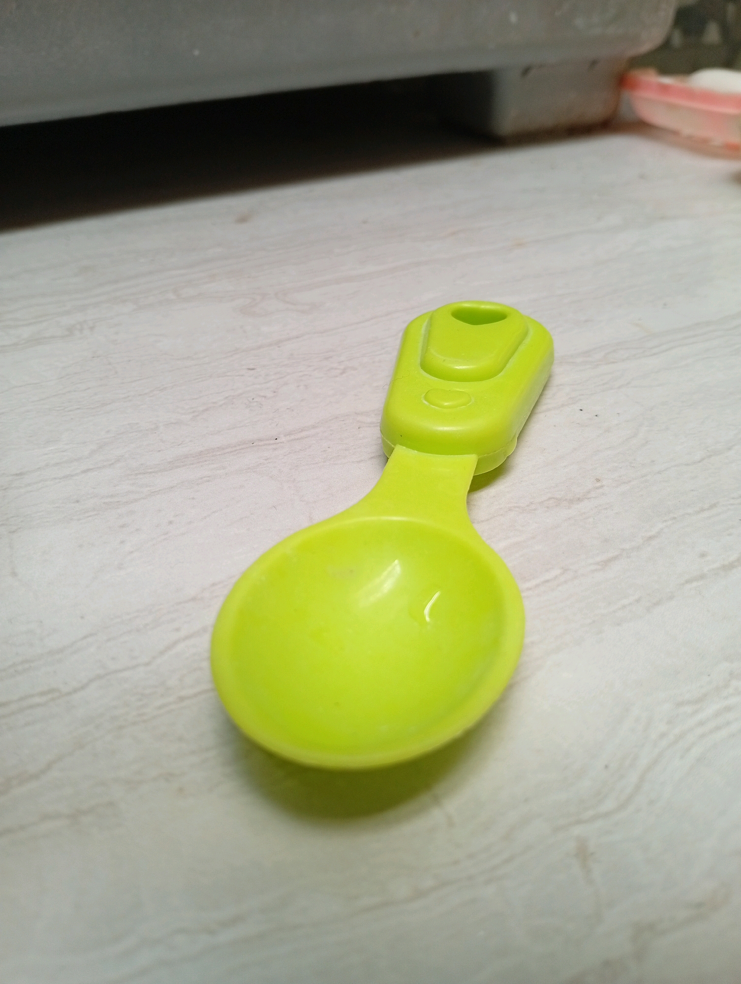 A single, bright green plastic measuring spoon lies on a light-colored, wood-patterned surface. The spoon is oriented diagonally, with the scoop end closer to the viewer and slightly to the left, and the handle extending upwards and to the right. The scoop of the spoon is slightly concave, and a small amount of clear liquid is visible within it. The handle is flat and has a small, rounded opening at the end. The surface it rests on has a texture resembling light-colored wood grain. In the upper portion of the image, a blurred, dark gray object is visible, casting a shadow on the surface below. The lighting appears to be overhead, creating a soft shadow beneath the spoon. The scene is indoors, on what appears to be a countertop or table.