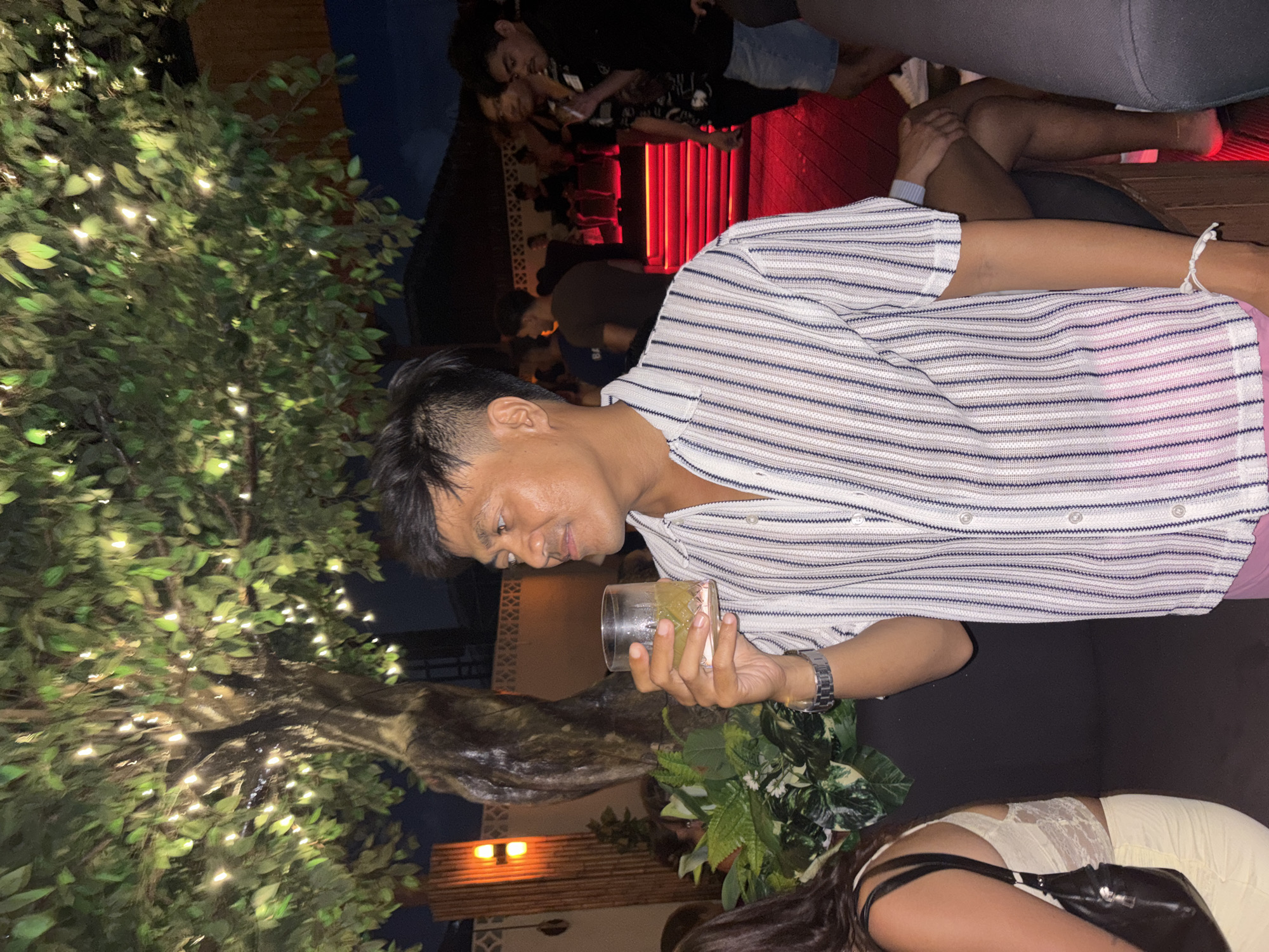This rotated image captures a vibrant night scene at an outdoor bar or lounge, likely in North Kuta, Indonesia. The main subject, positioned in the foreground, is a man with short dark hair, wearing a white and light blue striped short-sleeved button-up shirt. He holds a clear glass containing a light-colored beverage, possibly a cocktail, and looks towards the right with a calm, slightly pensive expression. A watch is visible on his left wrist.
To the man's left, a large artificial or decorative tree, adorned with numerous small, warm white string lights, creates a soft, inviting glow. Dark seating or cushions are visible, suggesting a comfortable lounge setting. In the lower right corner, parts of another person are visible, including a back and shoulder with long dark hair, wearing a light-colored top with lace details and carrying a black shoulder bag. Additionally, the crossed legs of at least two other individuals are visible, with one arm displaying a white beaded bracelet.
The background reveals more patrons socializing. A notable feature is a section of vertical red lights or panels, casting a distinct red glow across the area. Other warm ambient lights are scattered throughout the background, illuminating the faces and figures of more people. The dark sky confirms the setting is during the night or late evening. The scene exudes a relaxed and social atmosphere, characteristic of a nightlife venue. There is no visible text in the image.