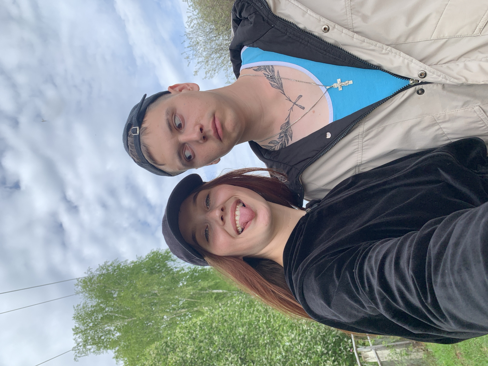 Two people are in the frame. The person in the foreground has reddish-brown hair, a black velvet-like jacket, and a black cap. They are sticking out their tongue and smiling. Behind and above them is another person wearing a blue shirt, a beige and black jacket, and a black cap. They have a tattoo on their neck and are wearing a silver chain with a cross pendant. The background shows trees and a cloudy sky. The image appears to be taken outdoors, possibly in a park or green space. The location within Фоки, Russia, cannot be definitively determined without additional information or landmarks.