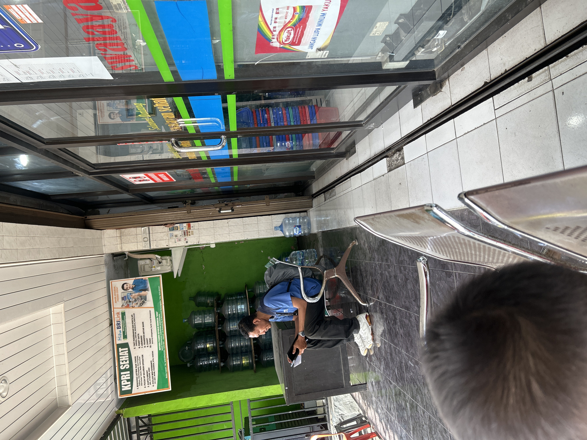 The image depicts an interior view of a health clinic or a similar facility in Wonosari, Indonesia. In the foreground, a person is seated on a metal chair, looking down at a dark-colored tablet. They are wearing a blue short-sleeved shirt, black trousers, and white sneakers. A dark backpack is visible on the chair next to them. The floor is tiled with dark gray squares.
To the left of the seating area, there is a dark wooden counter with several large plastic water jugs stacked on shelves behind it. A bright green wall is visible behind the water jugs, with a metal grate on the far left. A sign with text and images, including a BRI Link logo and a picture of a man, is mounted on the green wall.
The right side of the image features a glass-fronted storefront with blue and green horizontal stripes. Through the glass, stacks of colorful plastic baskets and more water jugs are visible. A sign with a Yakult advertisement, featuring text in Indonesian, is affixed to the glass. Another sign with text and red circular icons is also visible on the exterior of the glass. The overall environment suggests a commercial establishment that likely offers health services and potentially related products. The lighting is bright, indicating it is daytime.