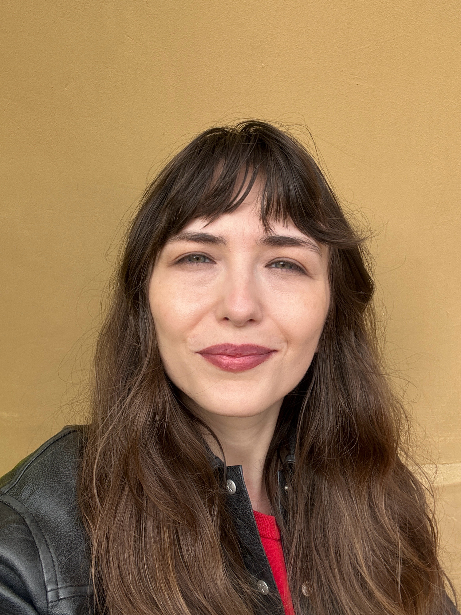 A medium shot photograph captures a woman with long, dark brown hair and bangs, wearing a black leather jacket over a red top. She is looking directly at the camera with a slight smile. The background is a plain, textured, ochre-colored wall. The image appears to be taken outdoors, possibly in Saint Petersburg, Russia, given the context provided, though no specific landmarks are visible to confirm this location. The lighting is soft and even, suggesting natural daylight.
