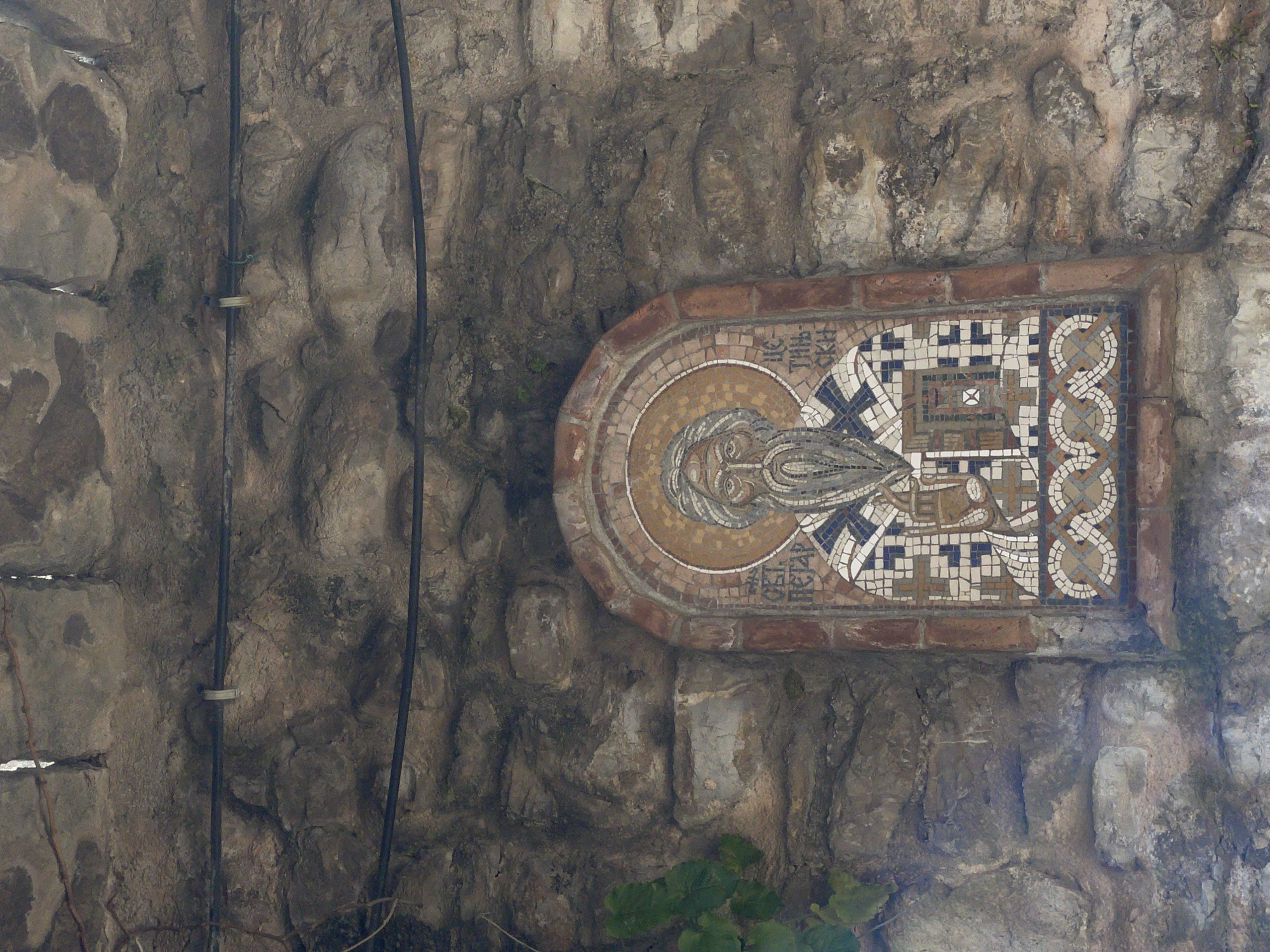 A mosaic is embedded in a rock wall. The mosaic depicts a person with a beard, halo, and priestly vestments. An inscription in a non-Latin alphabet is visible above the person. The mosaic is bordered by bricks. Two black electrical wires are attached to the wall with a fastener. Green foliage is visible at the bottom of the wall. The city is unknown.