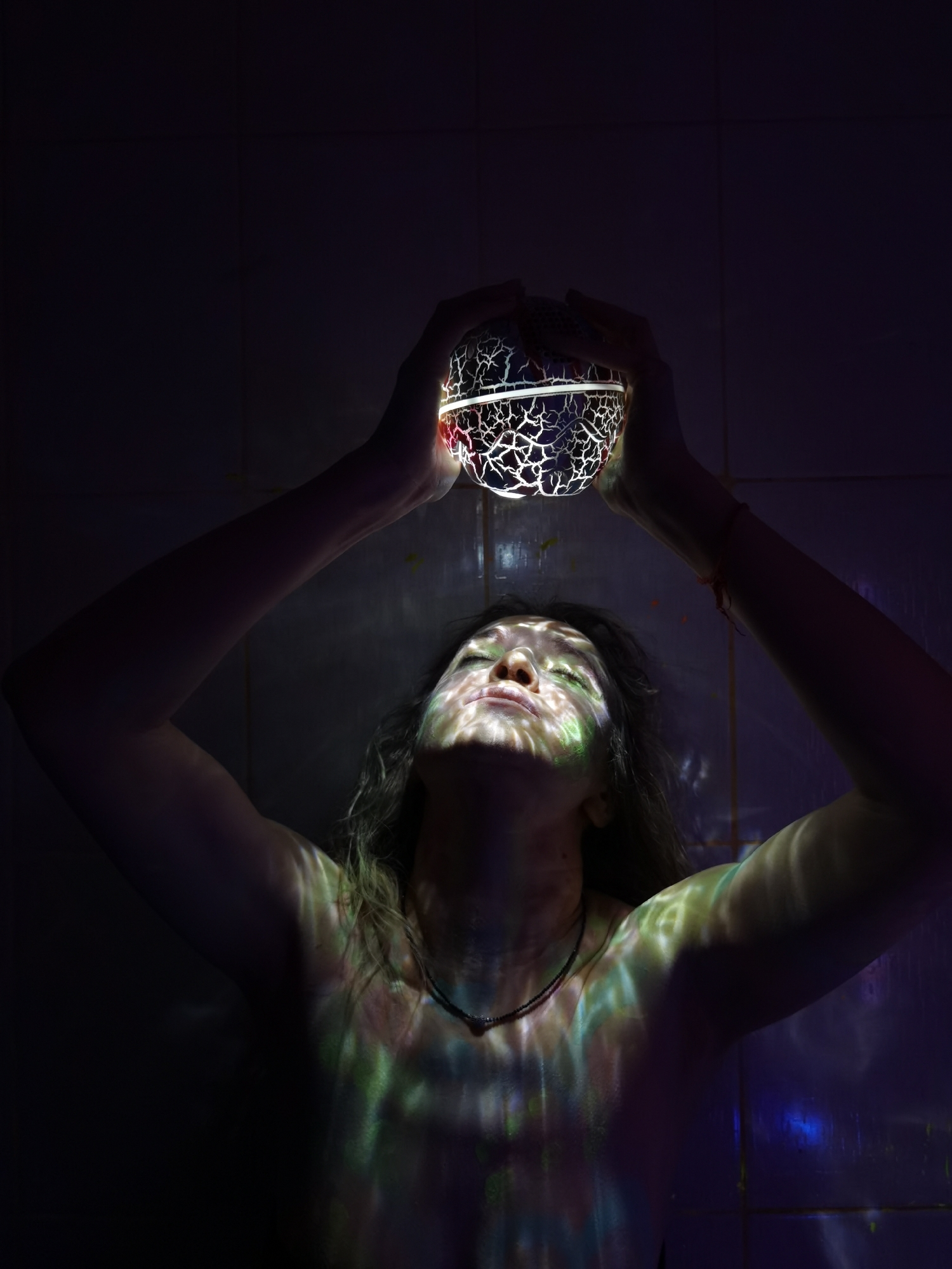 A vertical shot captures a person holding a glowing, patterned orb directly above their head in a dark indoor setting. The subject's arms are raised, with hands clasping the spherical object from underneath. The orb emits a bright, white-yellow light that highlights a distinct, cracked or shattered glass-like texture on its surface.
The person's head is tilted back, eyes closed, and their face is angled upwards, bathed in the light emanating from the orb. This light projects intricate, colorful patterns – primarily in shades of green, yellow, and purple – onto their face, neck, chest, and arms, creating a mesmerizing mosaic of light and shadow. Long, wavy hair is visible, cascading around the shoulders, and a simple, dark beaded necklace adorns the person's neck.
The background is mostly dark, suggesting a low-light environment. Faint outlines of what appear to be tiled walls can be seen in the deeper shadows, indicating an indoor location. The overall mood is contemplative, mystical, and serene, focusing on the interplay of light and form. There is no visible text in the image.