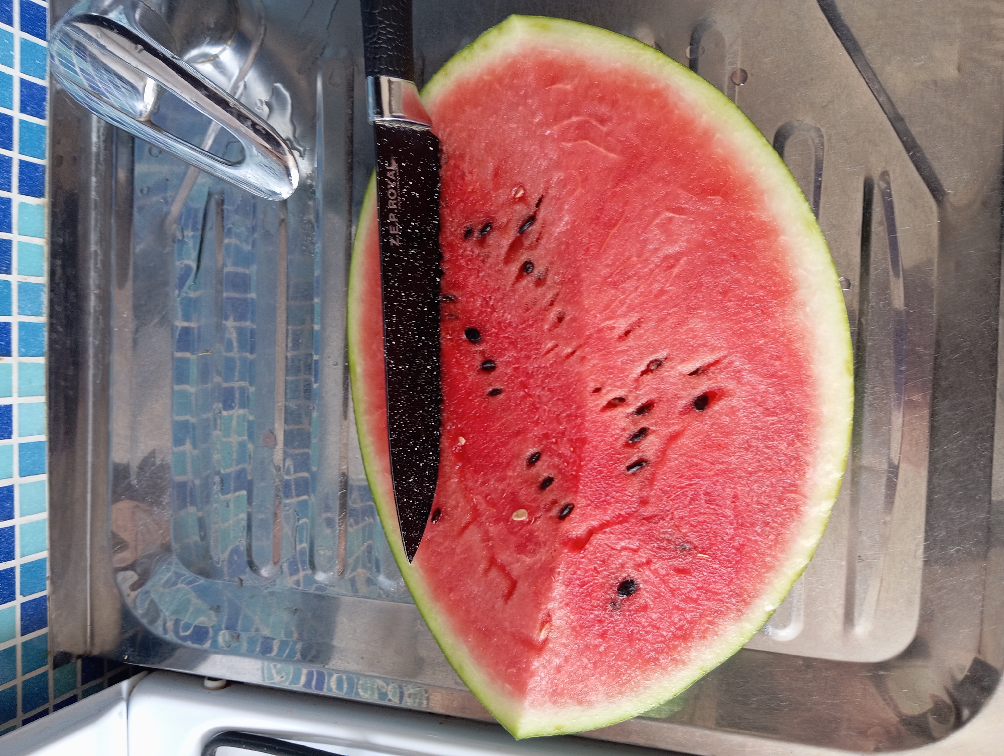 A close-up, overhead shot shows a slice of watermelon on a stainless steel sink. A black knife with a speckled blade rests on the watermelon, partially slicing into it. The watermelon is bright red with black seeds. The sink has a textured surface and a chrome faucet is visible in the background. The sink area is bordered by a wall tiled with blue and white squares. The scene is well-lit, likely by natural light. This image is not representative of any location in Temryuk, Russia, as it depicts a domestic kitchen scene.