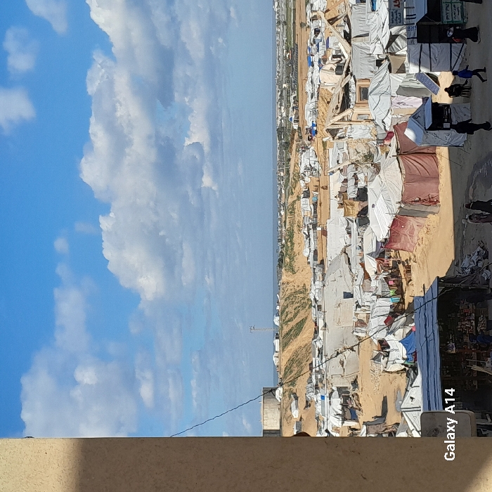 The image displays an expansive temporary settlement situated in Khan Yunis, Palestinian Territory, viewed from an elevated perspective. The scene is dominated by a dense arrangement of makeshift shelters and tents constructed from various materials including white, beige, and red-brown tarpaulins, fabrics, and plastic sheeting, often supported by visible wooden frames. The ground is primarily exposed, light brown sand or earth, with visible pathways winding through the camp.
In the immediate foreground, a beige wall partially obstructs the left side of the frame, from which a dark cable descends diagonally towards the lower-right. The "Galaxy A14" watermark is visible in the bottom-left corner. In the lower-midground, several individuals are observed walking along the sandy paths; approximately seven figures are discernable in the bottom-right, some appearing to wear head coverings. One structure in the bottom-left foreground functions as a market stall, displaying goods. Another structure in the mid-right exhibits significant damage, with exposed wooden beams forming a skeletal frame.
Beyond the immediate camp, a large, undulated sandy mound rises, featuring additional temporary shelters on its slopes. A large, cylindrical concrete water tank or silo stands prominently on the left side of this mound. To the right of the water tank, a tall, slender metal tower, possibly a communication or utility mast, is visible on the crest of the hill. In the far background, the landscape flattens to reveal distant structures, including what appear to be agricultural greenhouses, and an urbanized area. The horizon line reveals a vast body of water, identifiable as the Mediterranean Sea, under a partly cloudy blue sky. White cumulus clouds are scattered across the sky, indicating daytime conditions.