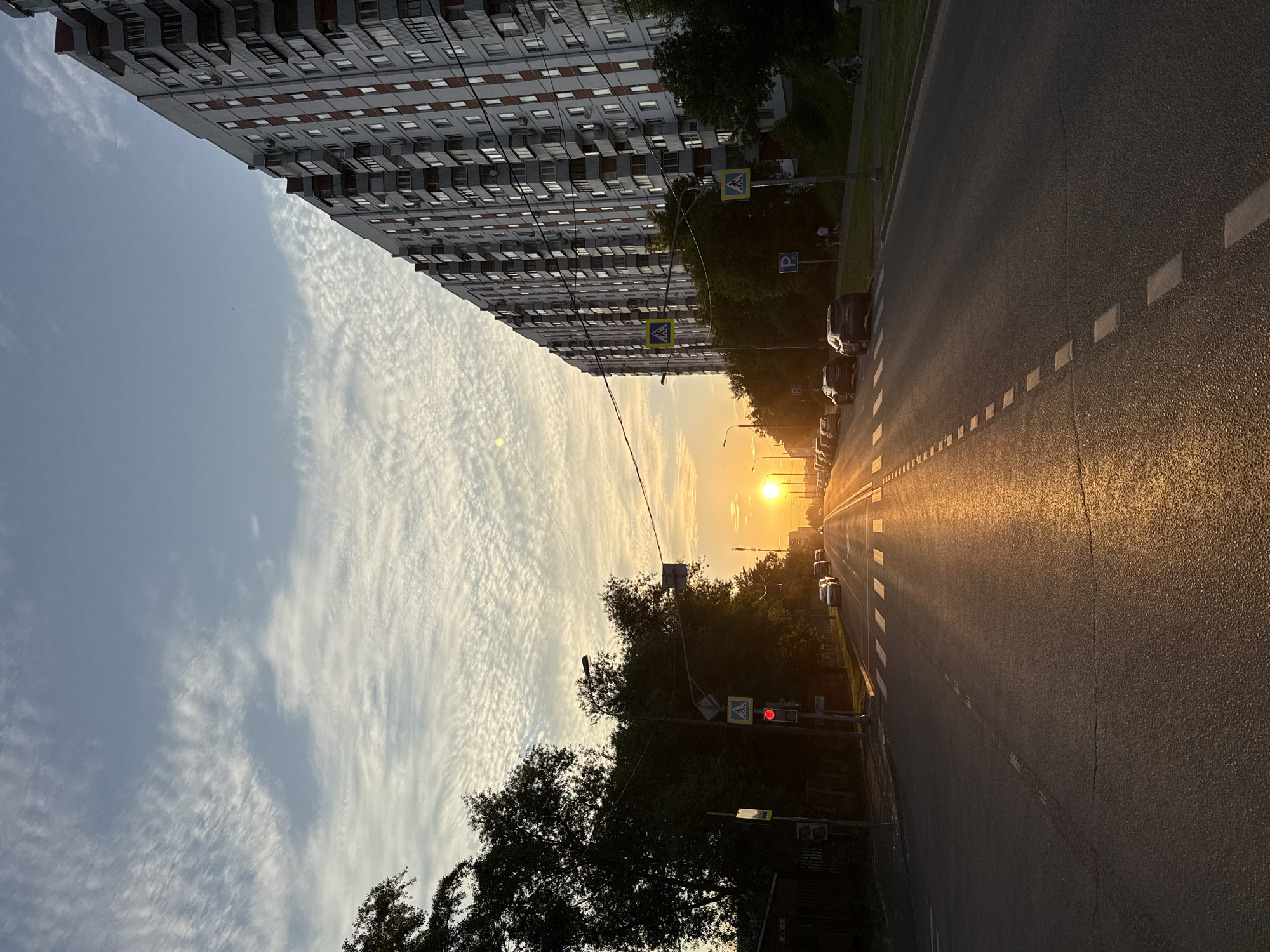 A street scene in Moscow, Russia, at sunset. The image is taken from a low angle, looking up towards a tall apartment building on the left and down a street lined with trees and parked cars on the right. The sun is setting on the horizon, casting a warm, golden glow over the scene and creating long shadows. The sky is filled with wispy clouds, and the light from the setting sun is reflected on the wet asphalt of the road.
The apartment building is a multi-story structure with many windows and balconies. Across the street, there are several cars parked along the side of the road. Pedestrian crossing signs are visible. A traffic light shows a red signal. The overall mood of the image is peaceful and serene, with a sense of the day winding down. The time of day is clearly indicated by the low sun and the long shadows. The weather appears to be clear, with some scattered clouds.
No visible text is discernible in the image.