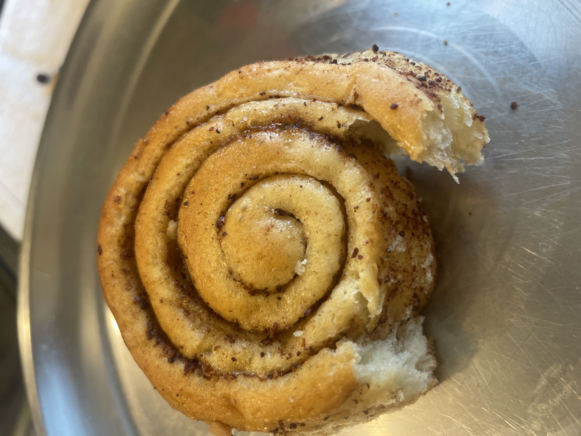 A close-up, overhead shot shows a single, partially eaten cinnamon roll on a metallic, circular plate. The roll is golden brown and has visible cinnamon swirls. A bite has been taken out of the left side of the roll, revealing its soft, white interior. The plate is reflective and appears to have some surface scratches. The background is blurred, but suggests an indoor setting with some natural light. The image does not contain any information about the city of Bhagsu, India.