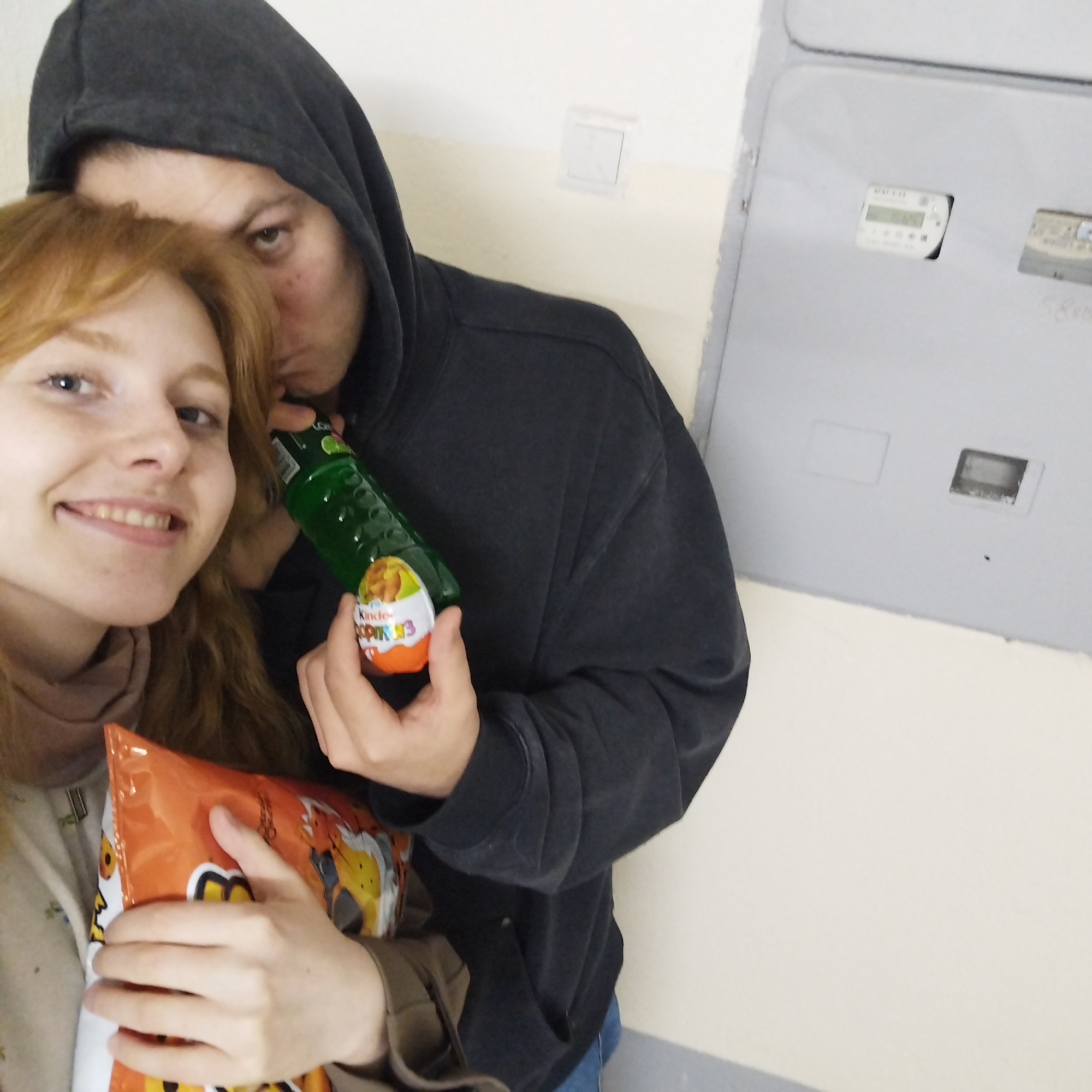 A close-up selfie shows two people, a woman and a man, indoors. The woman, with reddish-blonde hair, is in the foreground, smiling broadly at the camera. She holds a large orange bag of chips, which appears to be Cheetos due to the visible cartoon cheetah character. Behind her, a man wearing a dark hoodie is seen partially, with his face obscured as he appears to kiss the woman's head or hair. He holds a green, textured plastic bottle in one hand and a Kinder Surprise egg in the other, clearly labeled "Kinder Surprise Copines 3". The background features a plain light-colored wall and a large grey utility panel or box. This panel has at least two cutouts, revealing what appear to be electricity meters. One digital display shows numbers like "1325" and "1408", with a label at the top left that might read "MATUTO". The scene suggests a casual, affectionate moment, possibly taken in a residential hallway or utility area.