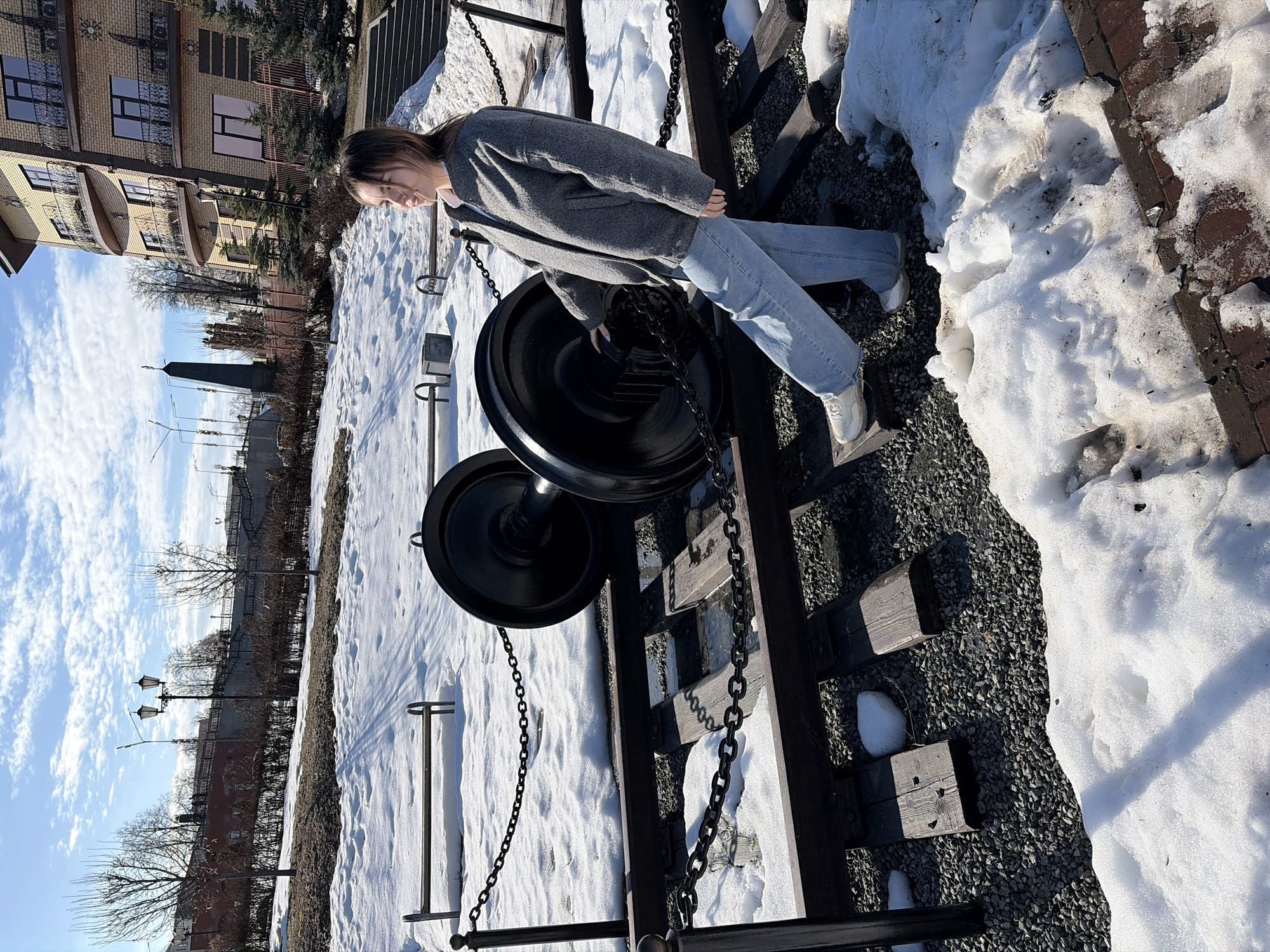 A young woman wearing a gray coat and light blue jeans stands beside a stationary black train wheel and chain apparatus, possibly a historical display, in Sarapul, Russia. The ground is covered in snow and gravel. Behind the woman is a brick building with balconies. A street with lamp posts and bare trees is visible in the background, as well as a tall monument-like structure. The sky is blue with scattered clouds.