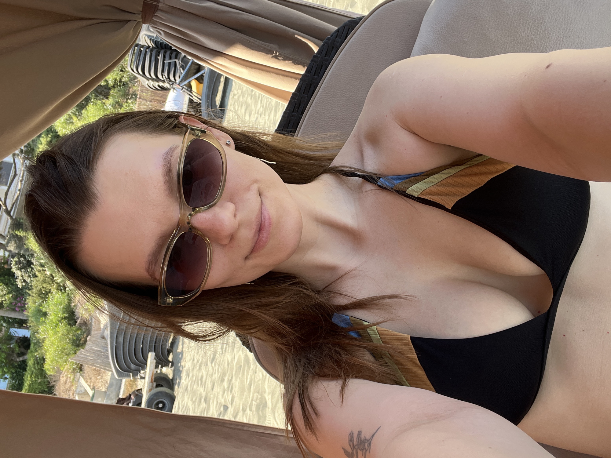 A medium shot shows a woman with brown hair and sunglasses on a beach in Agios Tychona Community, Cyprus. She is wearing a black bikini with a striped strap. She is seated in a covered lounge chair, with the beach and ocean visible in the background. There are stacks of chairs and a trailer visible on the sand. The woman is smiling and looking at the camera.