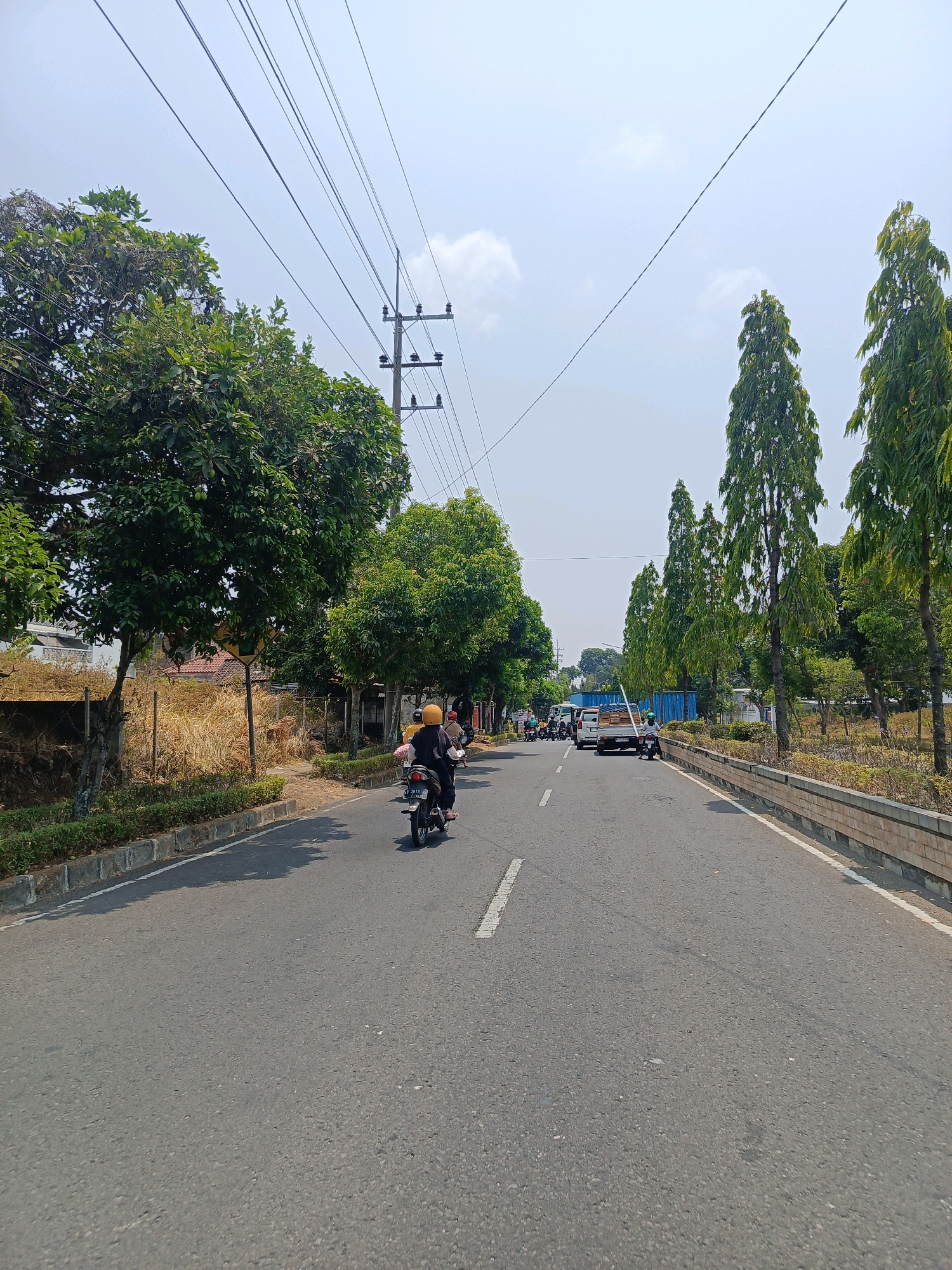 This image depicts a street scene in Sukowinangun, Indonesia. The road is asphalt and marked with white lines. Several vehicles are present, including motorcycles and cars. A person is riding a motorcycle in the foreground, wearing a yellow helmet. The sides of the road are lined with trees and vegetation, with some residential buildings visible in the background. Overhead, power lines stretch across the frame, supported by utility poles. The sky is visible and appears cloudy. The overall impression is a typical street in a populated area with moderate traffic.