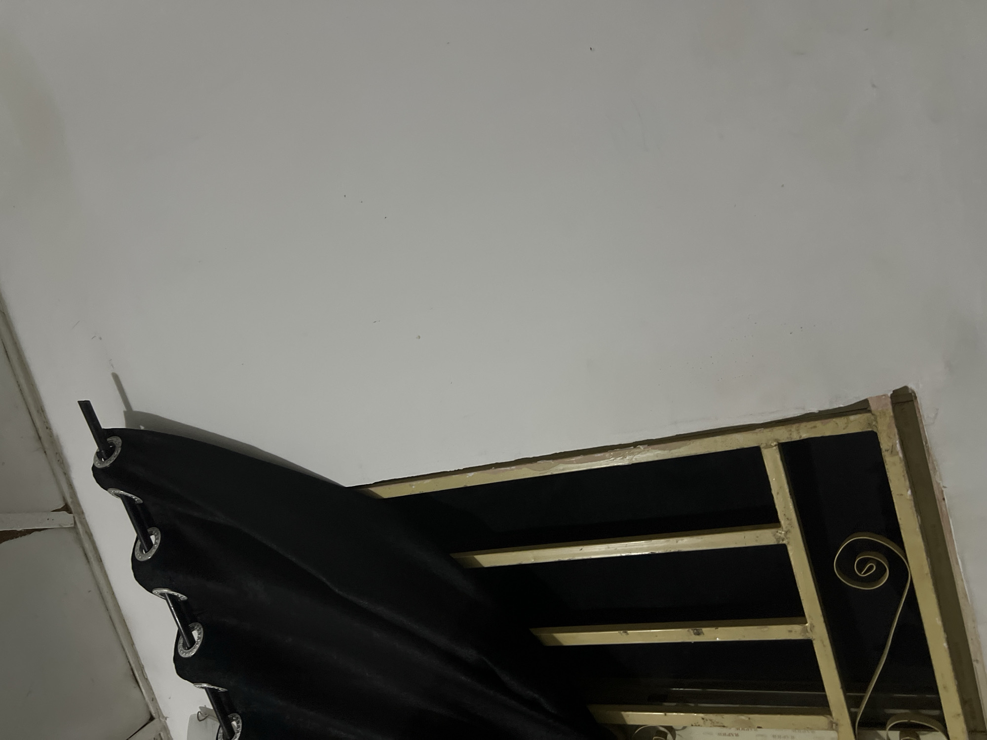 The image depicts a close-up view of a window with a black curtain partially drawn across it. The window features a yellow metal grill, with decorative scrollwork at the bottom. The curtain is held up by a dark metal rod, which extends across the top of the window. The wall behind the window is painted white and appears to be slightly textured. The ceiling above is also visible, with some signs of wear and tear. The lighting suggests it is either nighttime or the room is dimly lit. The scene is likely located indoors, possibly in a residential building. The image does not provide any specific clues to pinpoint its exact location within the city of Howayi, Nigeria, beyond the general context of an urban or semi-urban setting. There are no people or ongoing actions depicted in the image.